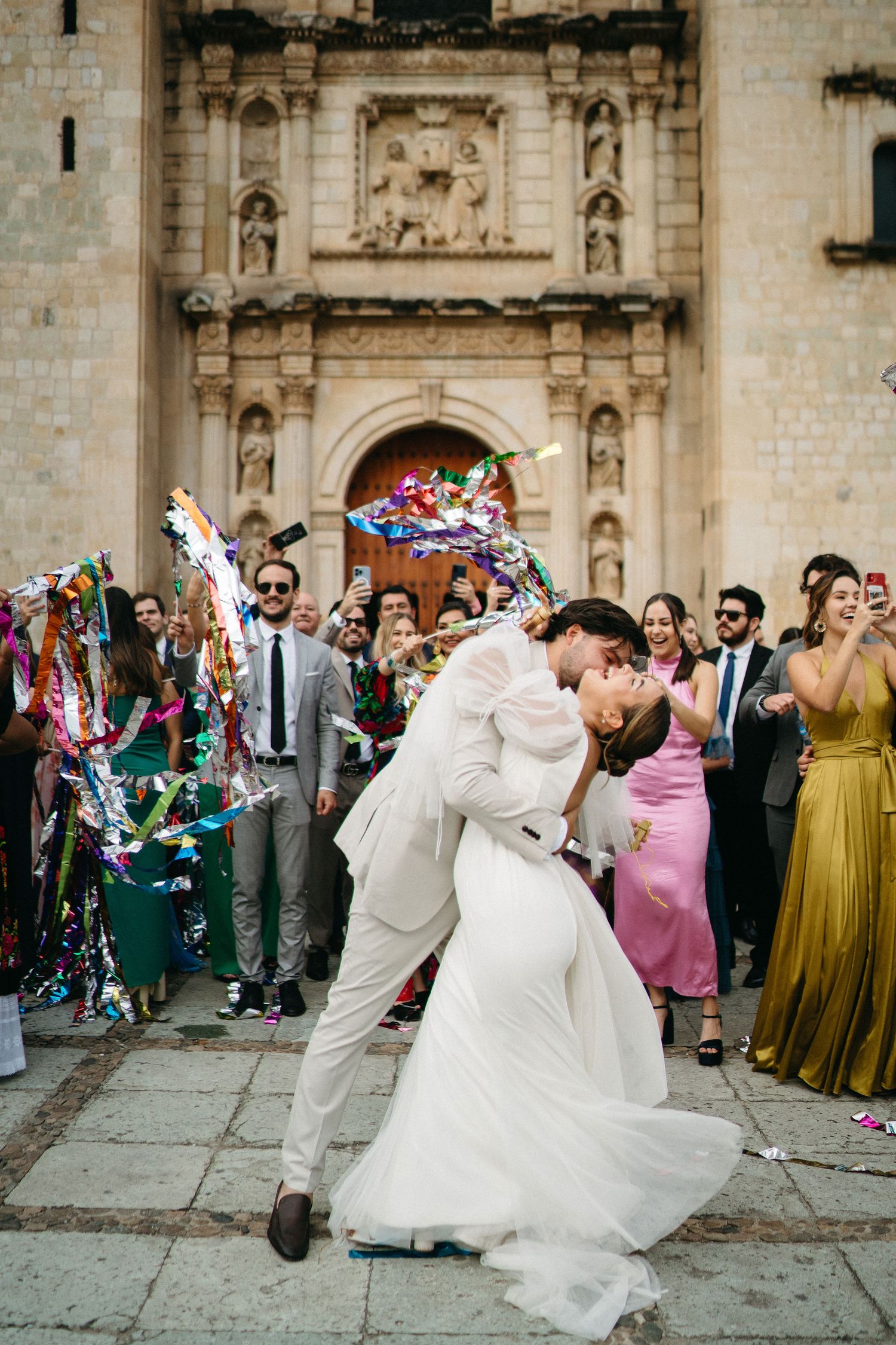 Wedding party celebrating with colorful streamers in front of an ornate stone church facade.