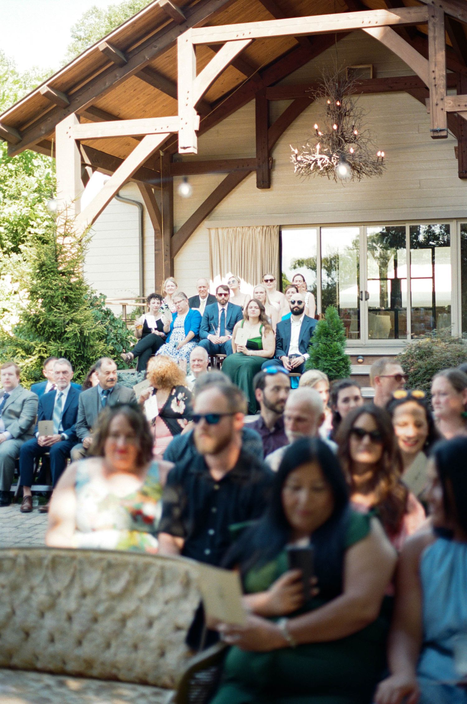 Wedding guests fill wooden benches under a covered outdoor pavilion.