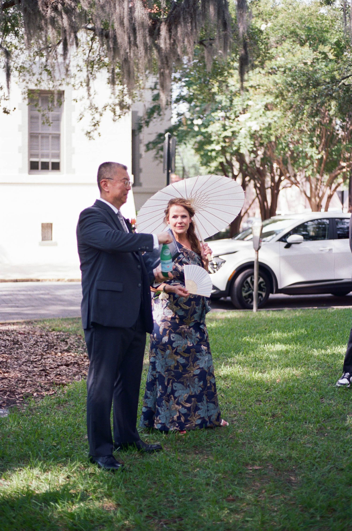 A couple poses under Spanish moss draped trees with a white parasol at an outdoor event.