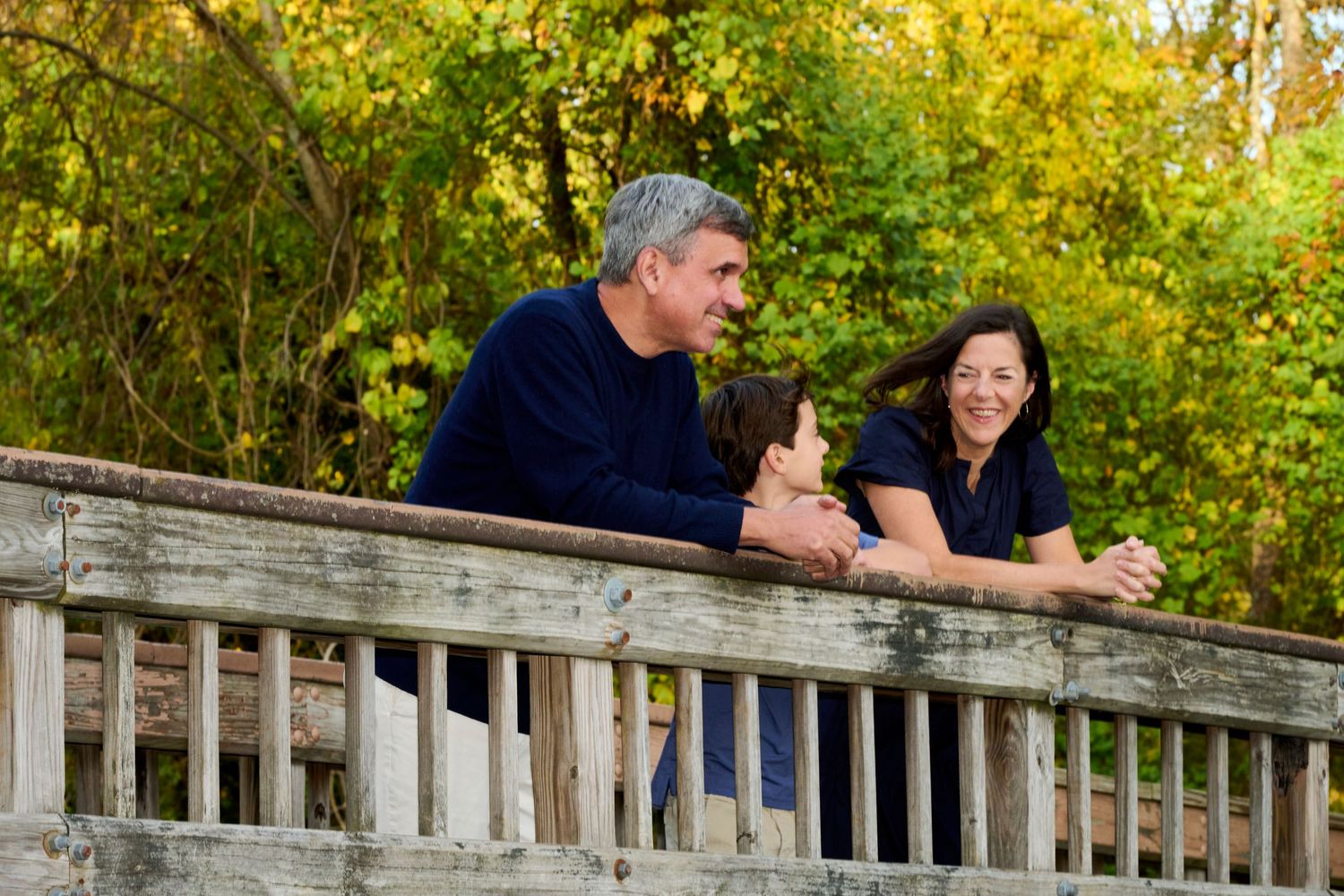 Family members sit on wooden railing with colorful fall trees behind them.