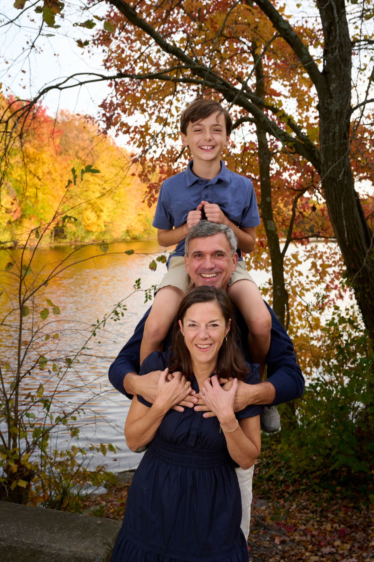 Family poses playfully together near lake with autumn trees in background.