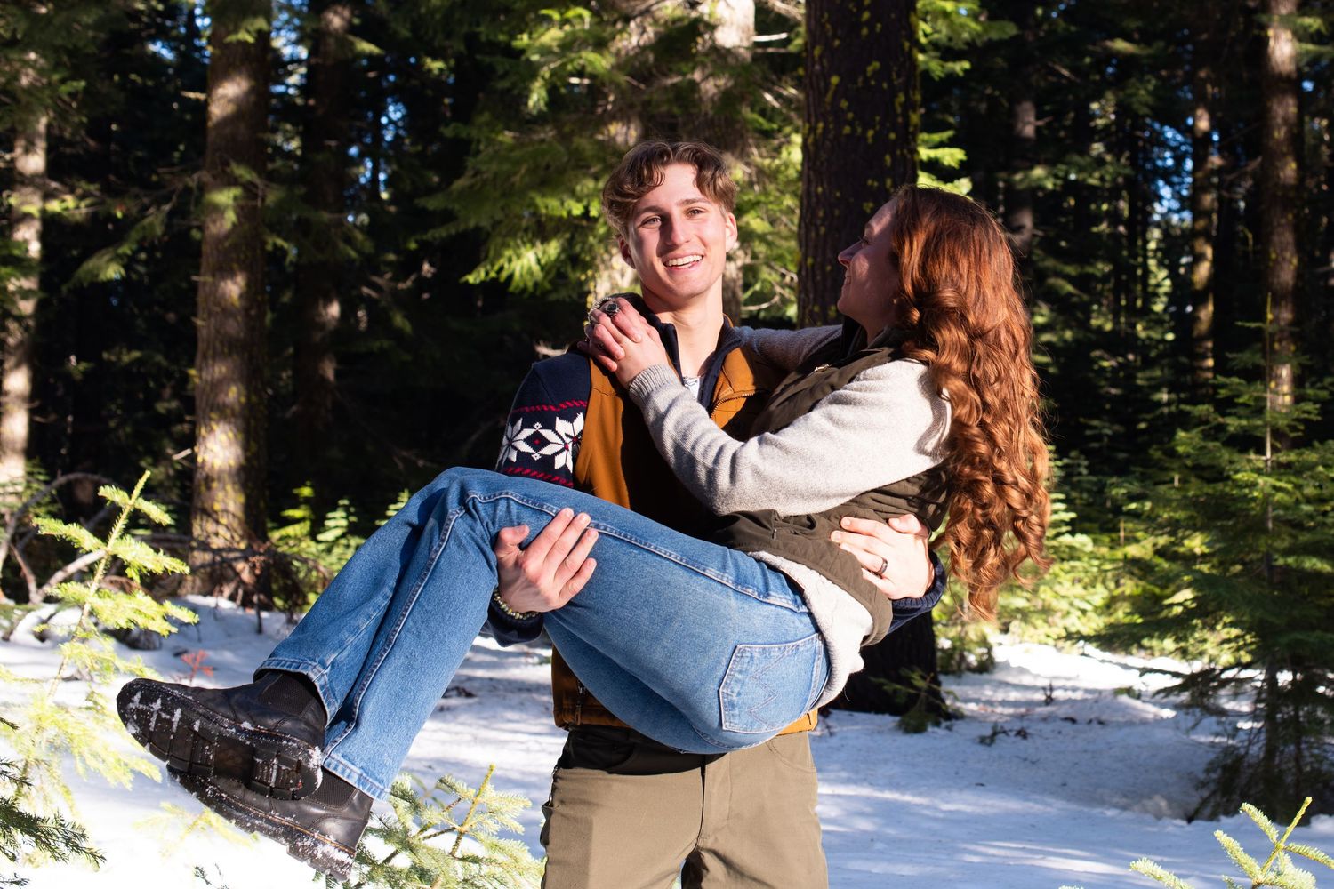 Man carrying smiling woman in his arms through a snowy pine forest on a sunny winter day.