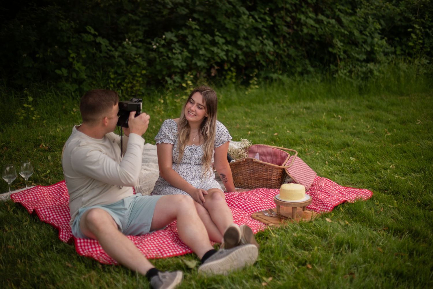 Man photographing his smiling girlfriend with a camera during a cozy outdoor picnic on a red blanket.