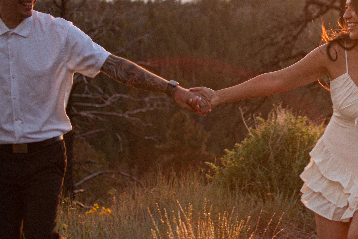 Couple holding hands at golden hour in a field, romantic outdoor engagement photo with warm sunset light.