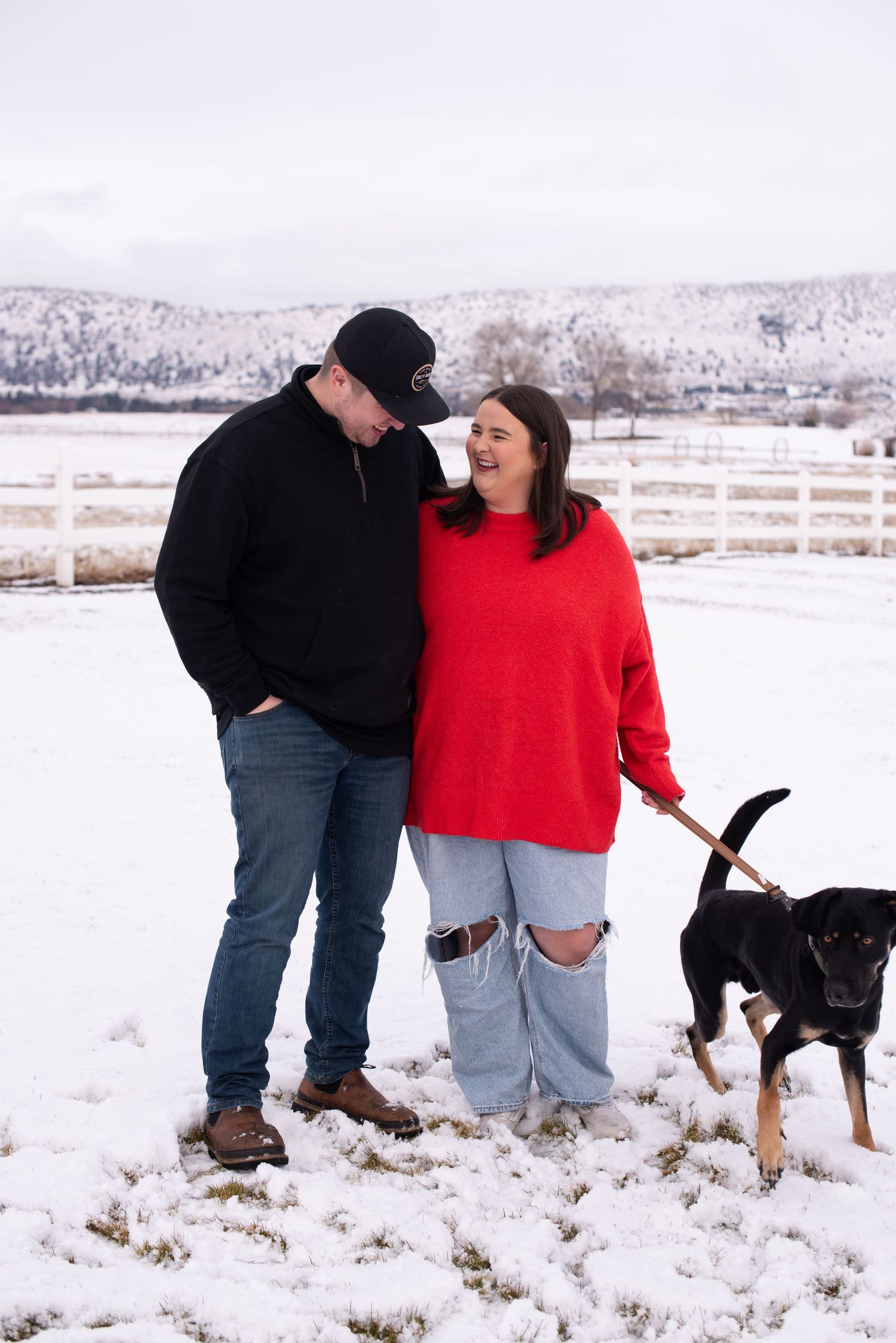 Couple in red and black sweaters smiling with their dog in a snowy ranch setting with white fence backdrop.