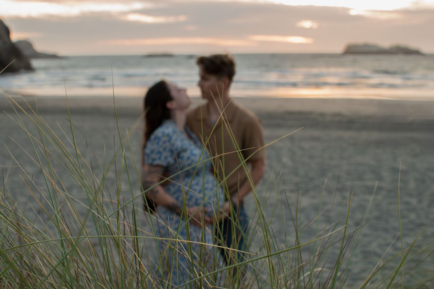 Couple sharing a tender moment on a beach at sunset, with tall grass in the foreground and ocean in the background.