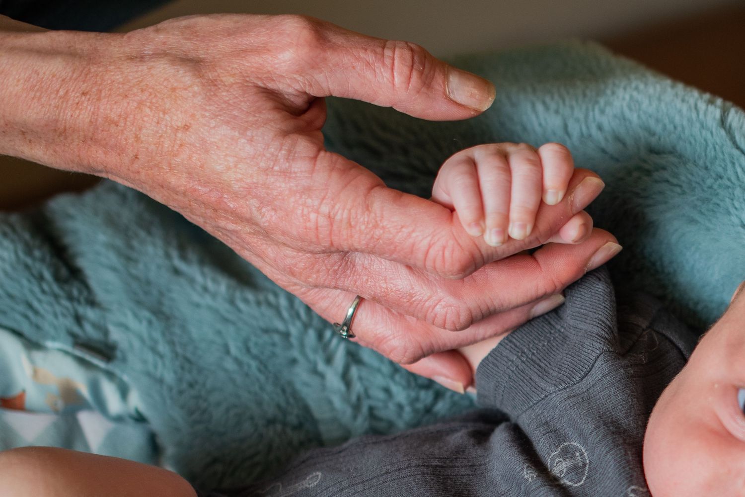 Elderly hand with ring gently holding a newborn baby's tiny fist on a teal blanket.