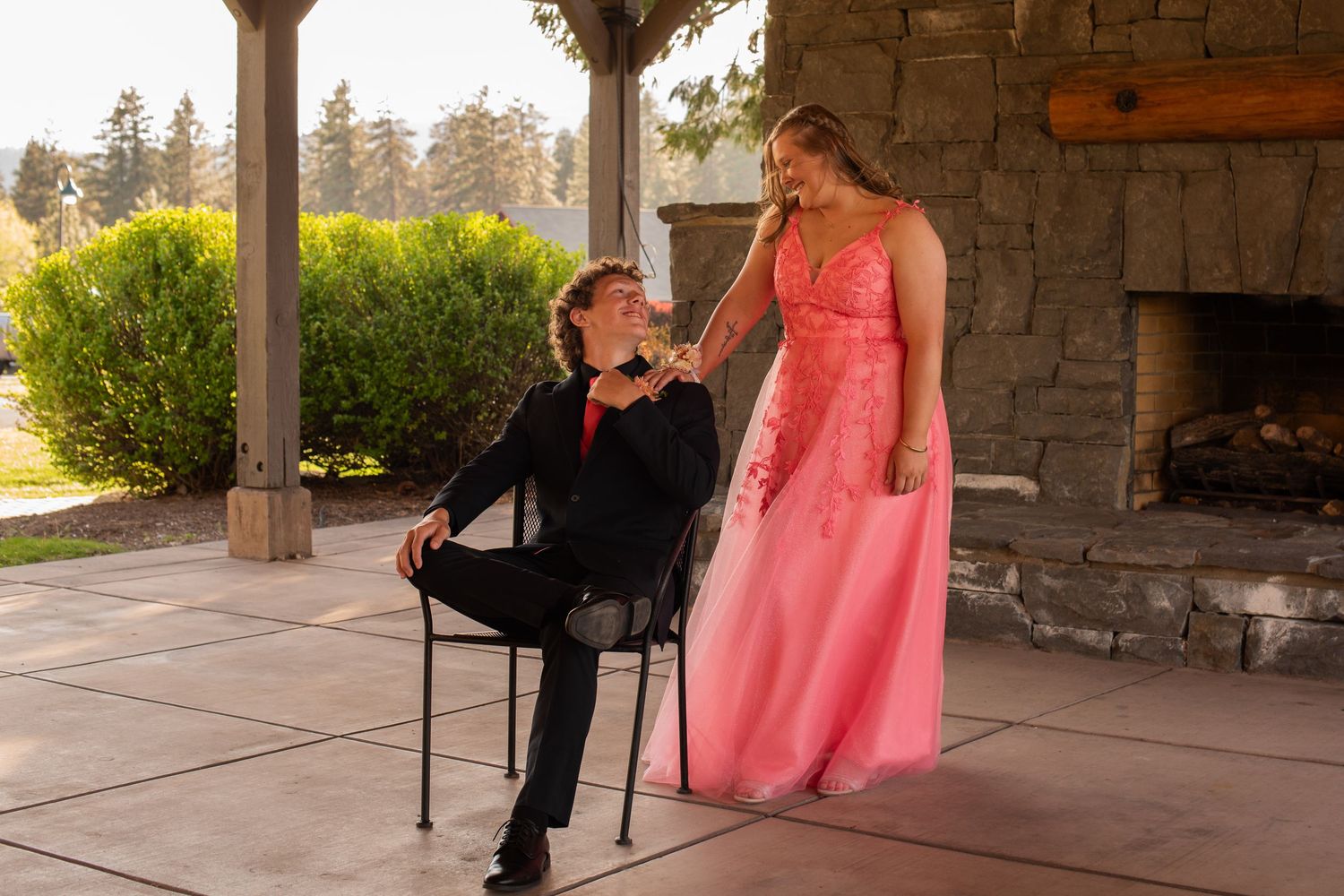 Teen couple in prom attire, boy in black suit seated, girl in pink gown standing outdoors by stone fireplace.