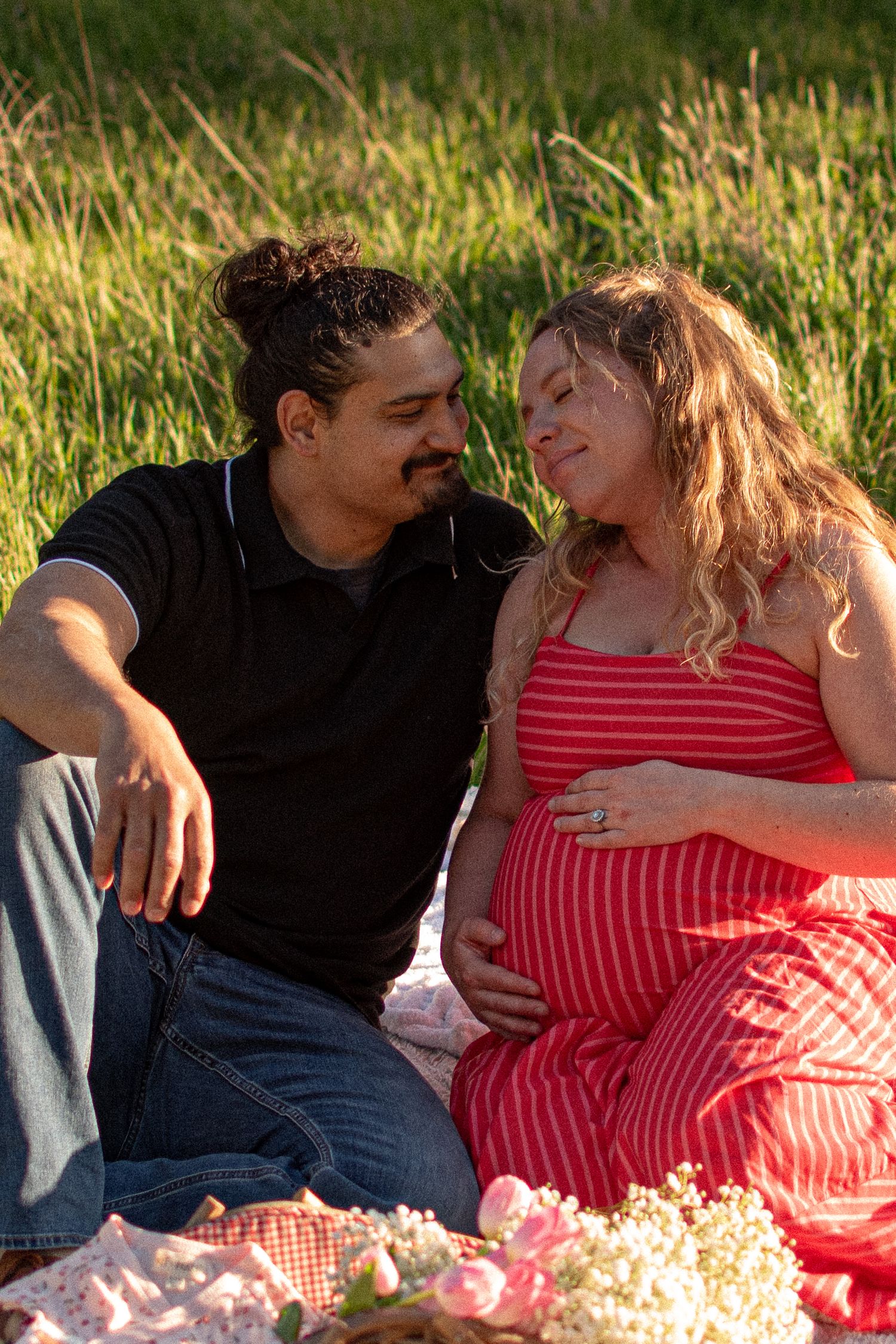 Pregnant woman in red striped dress sits with partner on picnic blanket in grassy field, cradling baby bump.
