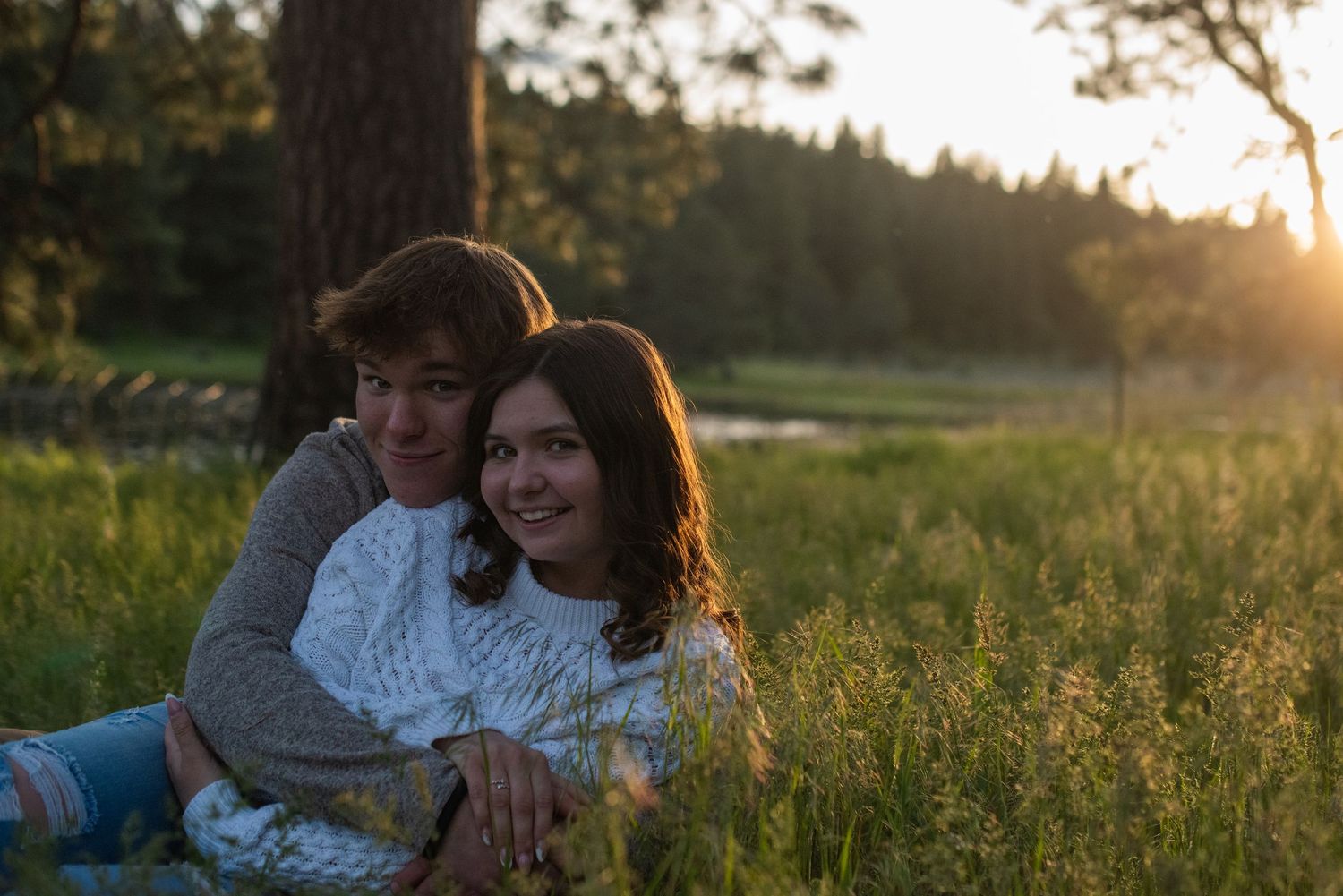 Couple sitting together in tall grass at sunset, smiling warmly in a pine forest.