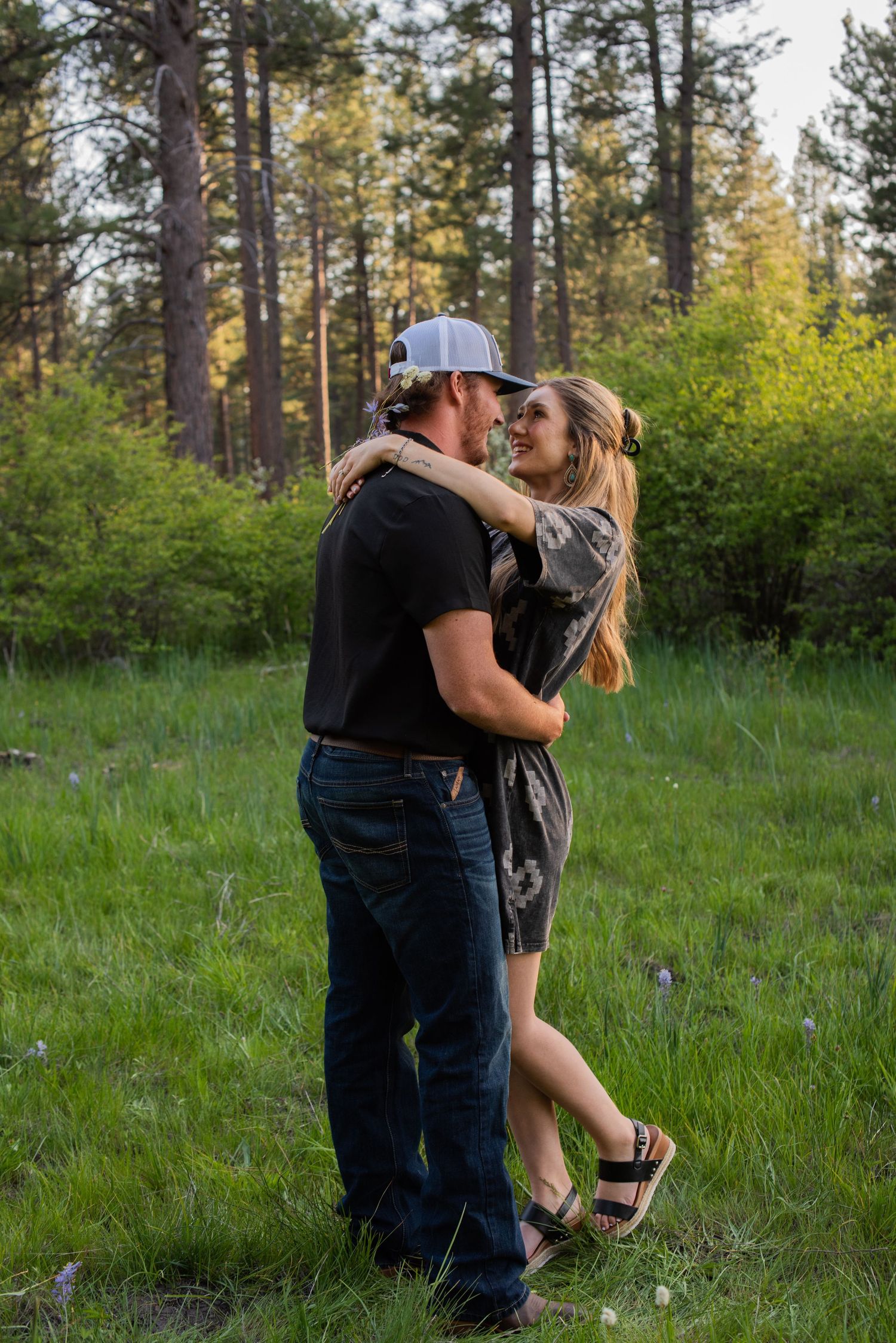 Couple embracing and smiling at each other in a lush green meadow surrounded by tall pine trees.