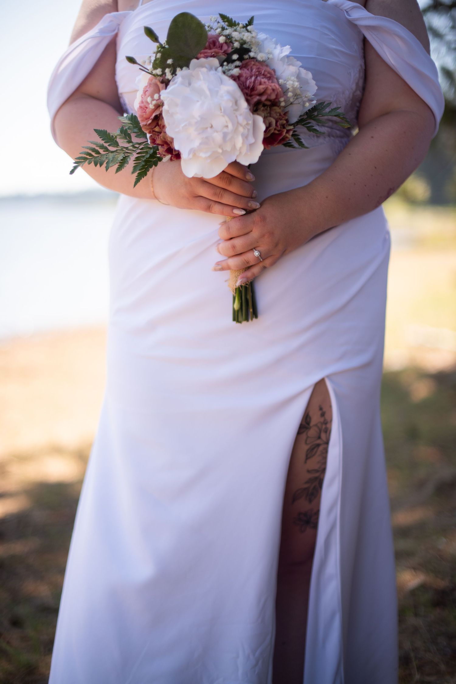 Close-up of bride in white off-shoulder gown holding a pink and white wedding bouquet with tattoo visible.