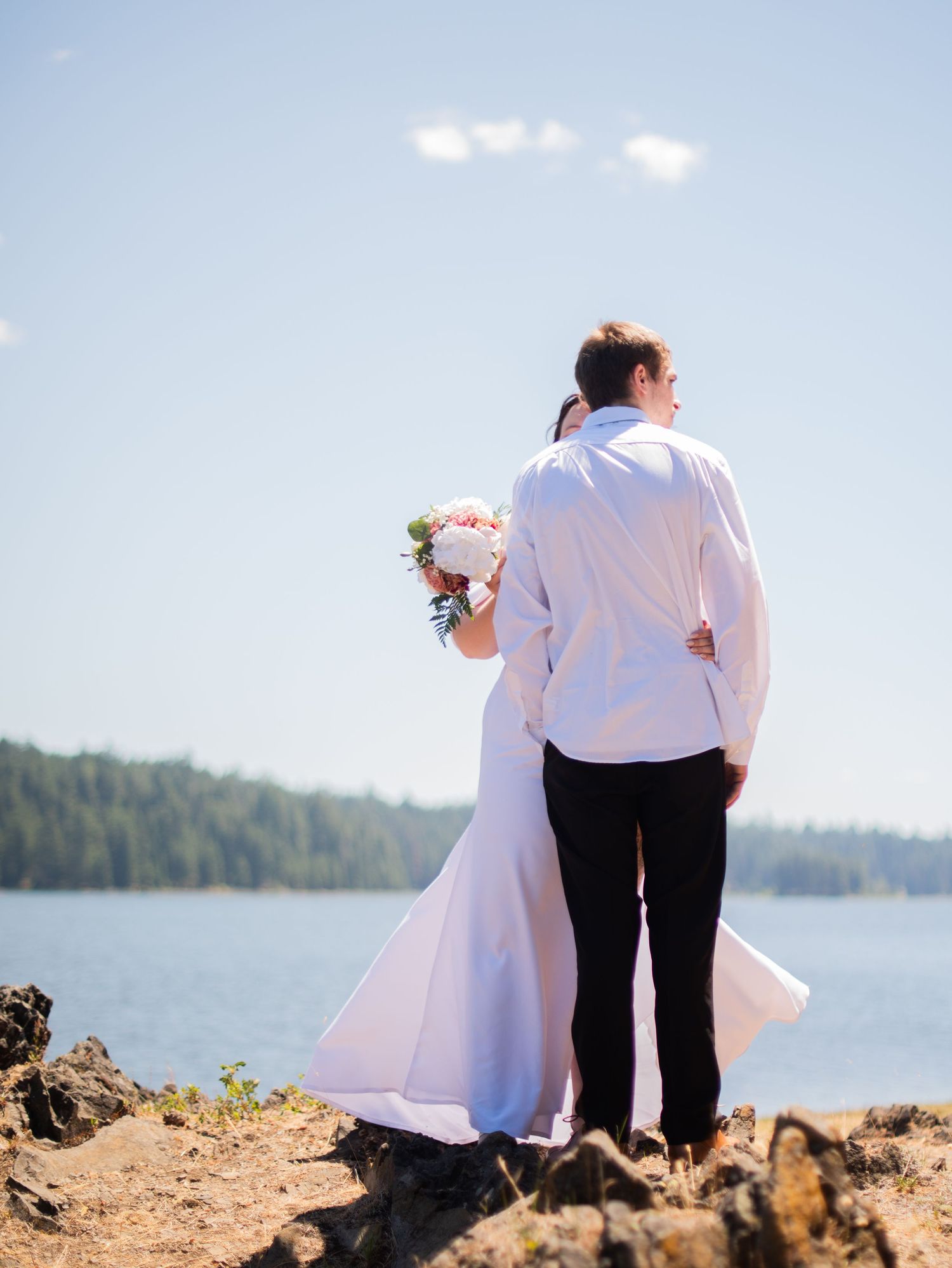 Bride and groom stand together overlooking a serene lake on a sunny wedding day.