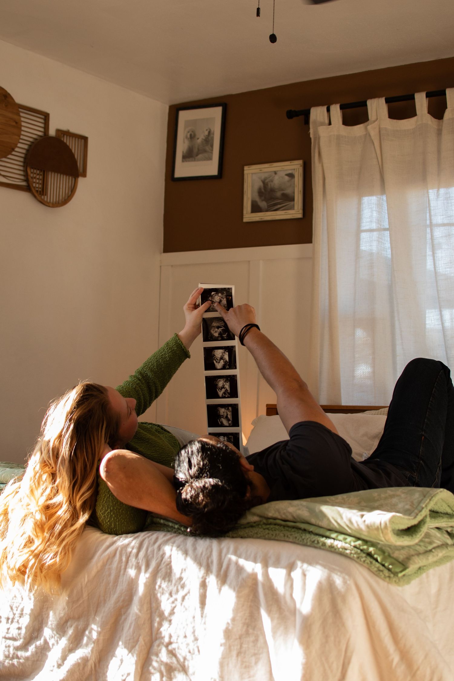 Couple lying on bed together looking at ultrasound photos in a sunlit bedroom with brown walls and white curtains.