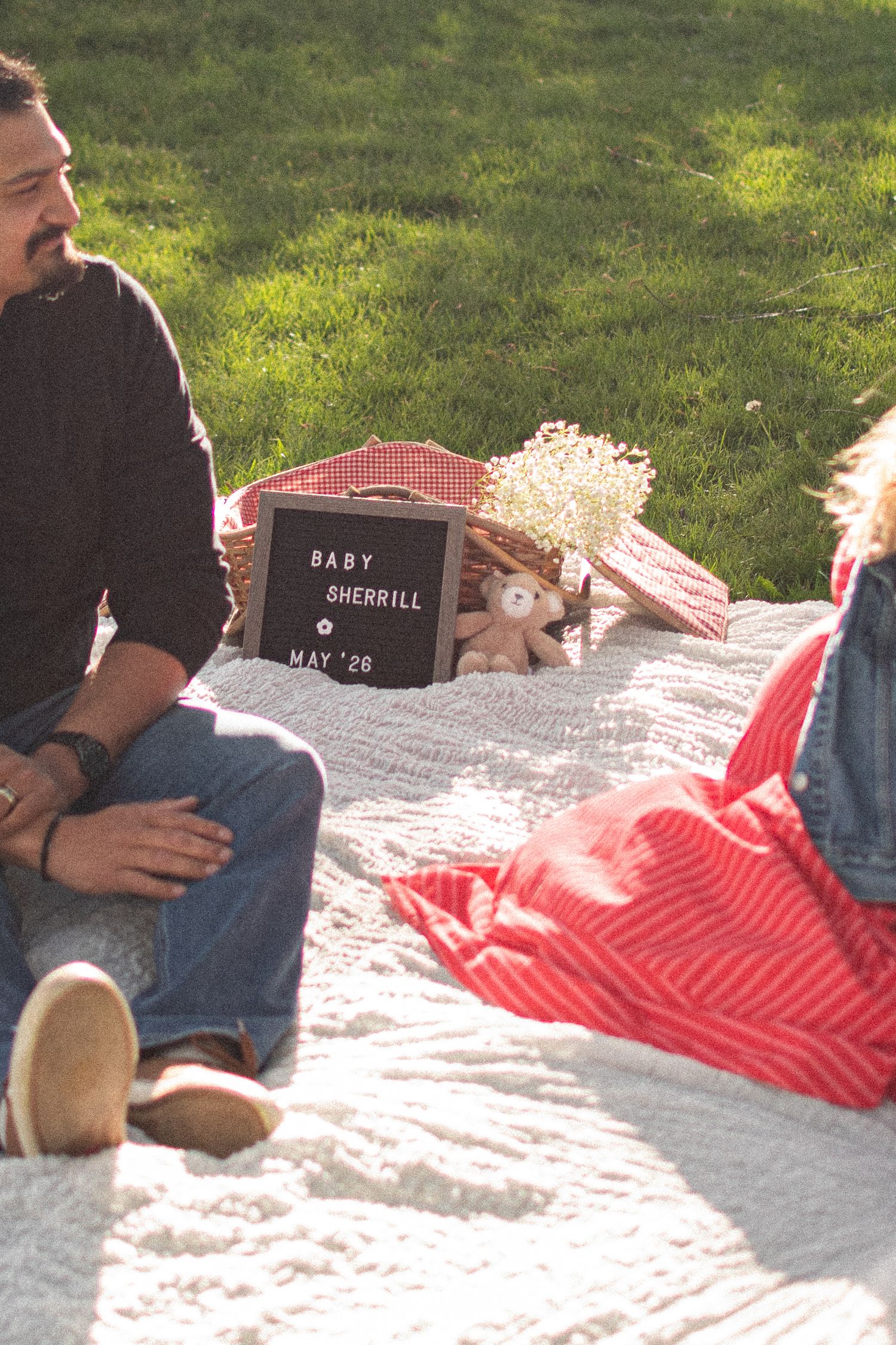 Couple sits on white blanket outdoors near a letter board sign reading Baby Sherrill May 26 with a stuffed animal.