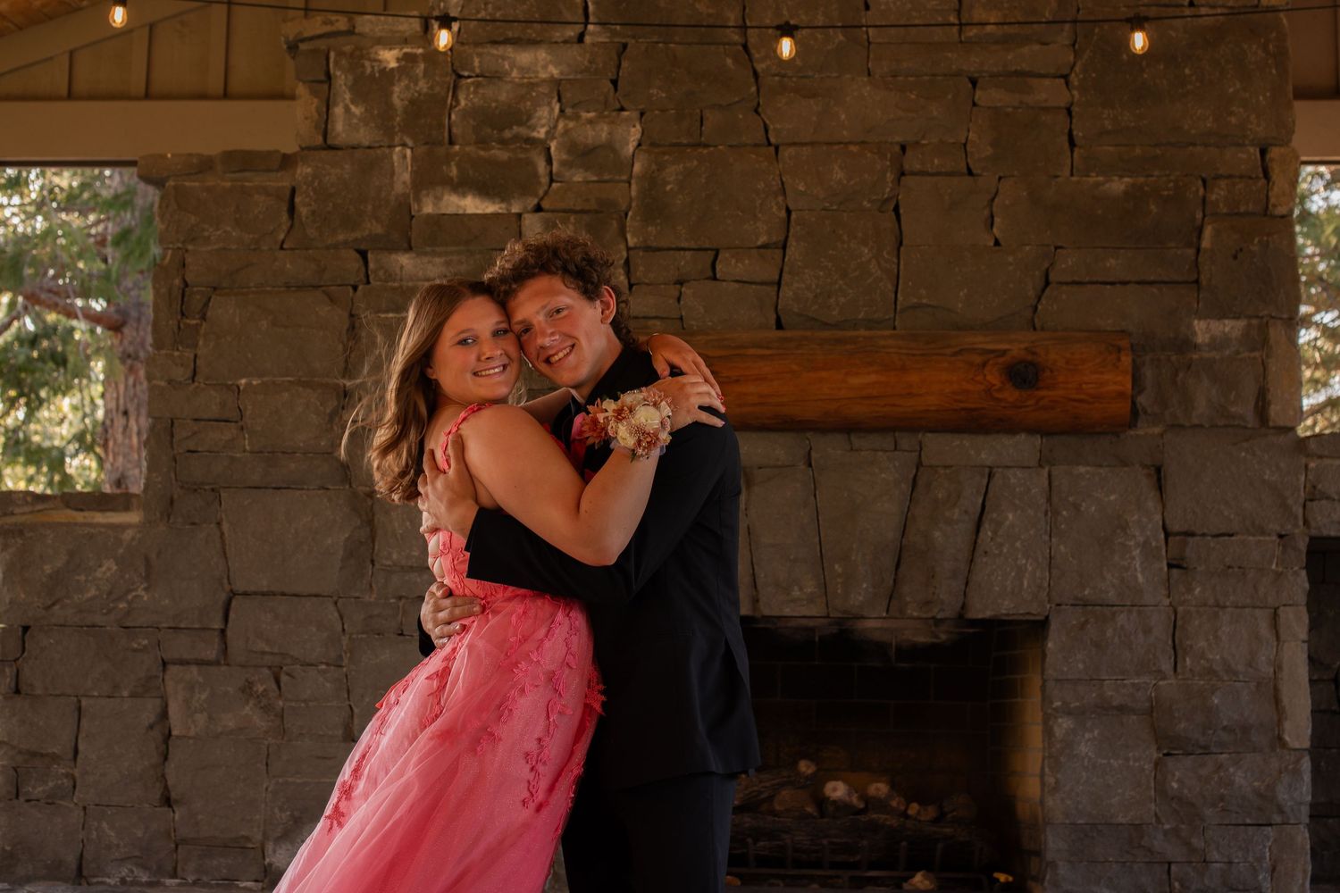 A couple in formal attire embraces romantically in front of a rustic stone fireplace at prom.