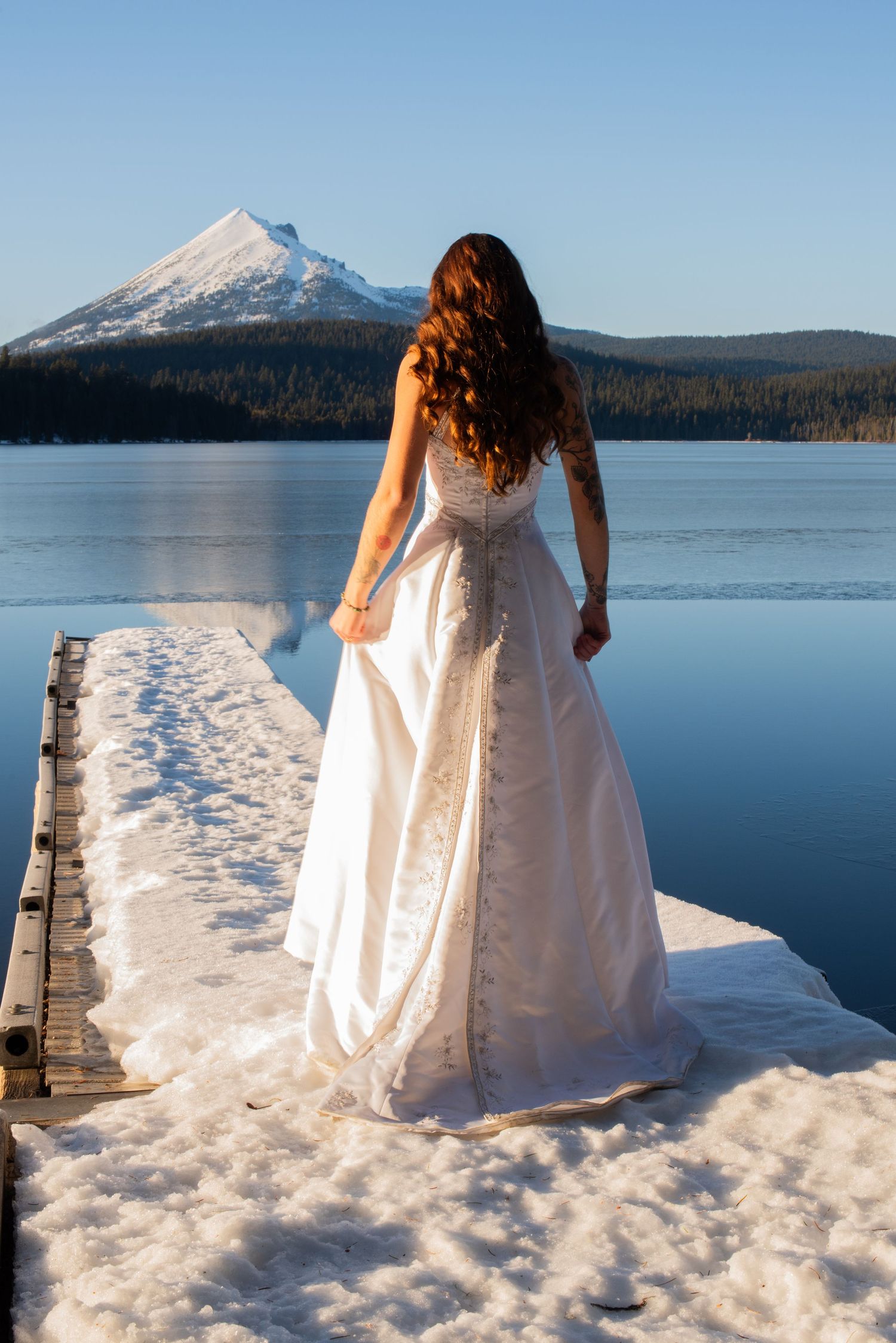 Bride in ornate white gown stands on a snow-covered dock overlooking a frozen mountain lake.