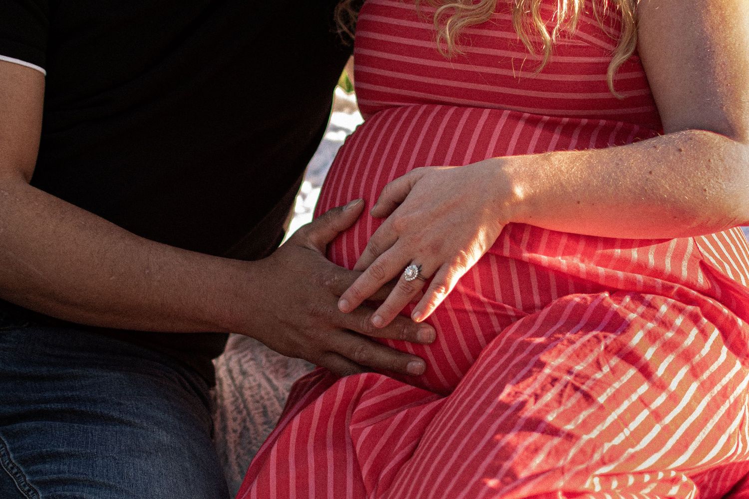 Couple sitting together, man placing hand on pregnant woman's belly in red striped dress, wearing engagement ring.