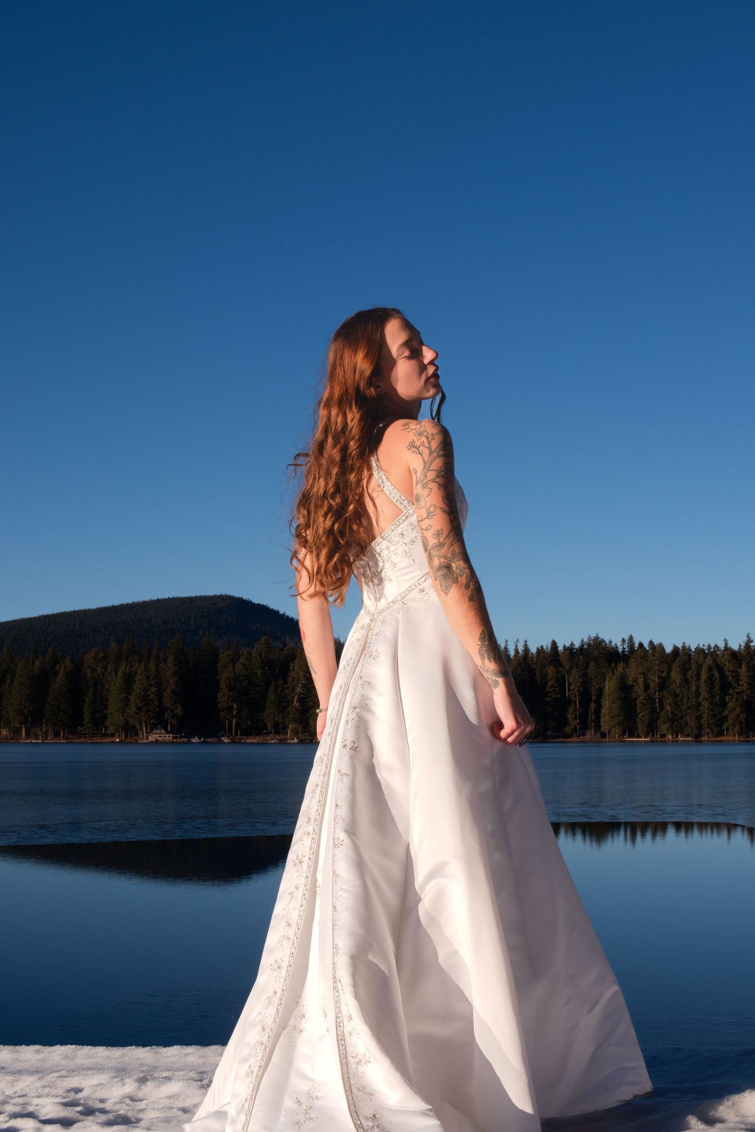 Tattooed bride in white wedding gown poses gracefully by a frozen lake under a clear blue sky.