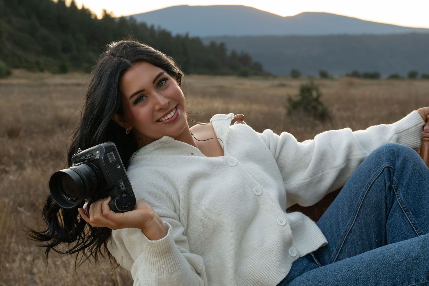 Smiling woman in white sweater and jeans holding a camera while relaxing in a golden field with mountains behind her.