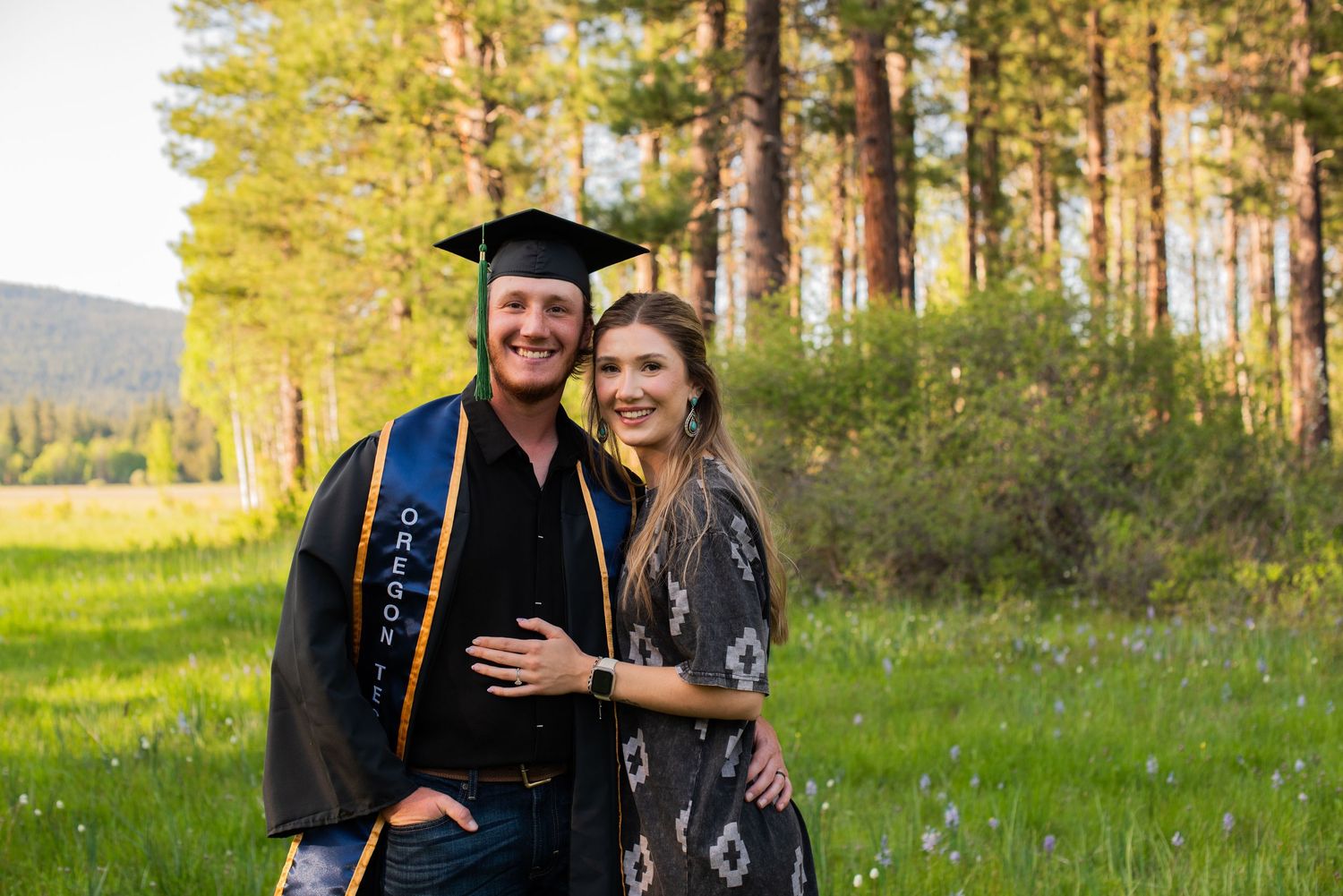 Graduate in cap and gown with blue stole posing with woman in green meadow with pine trees.