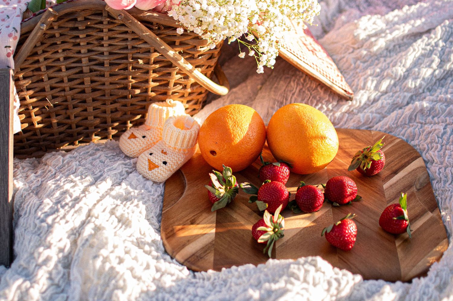 A rustic picnic spread with fresh oranges, strawberries, and cookies on a wooden board atop a white blanket.