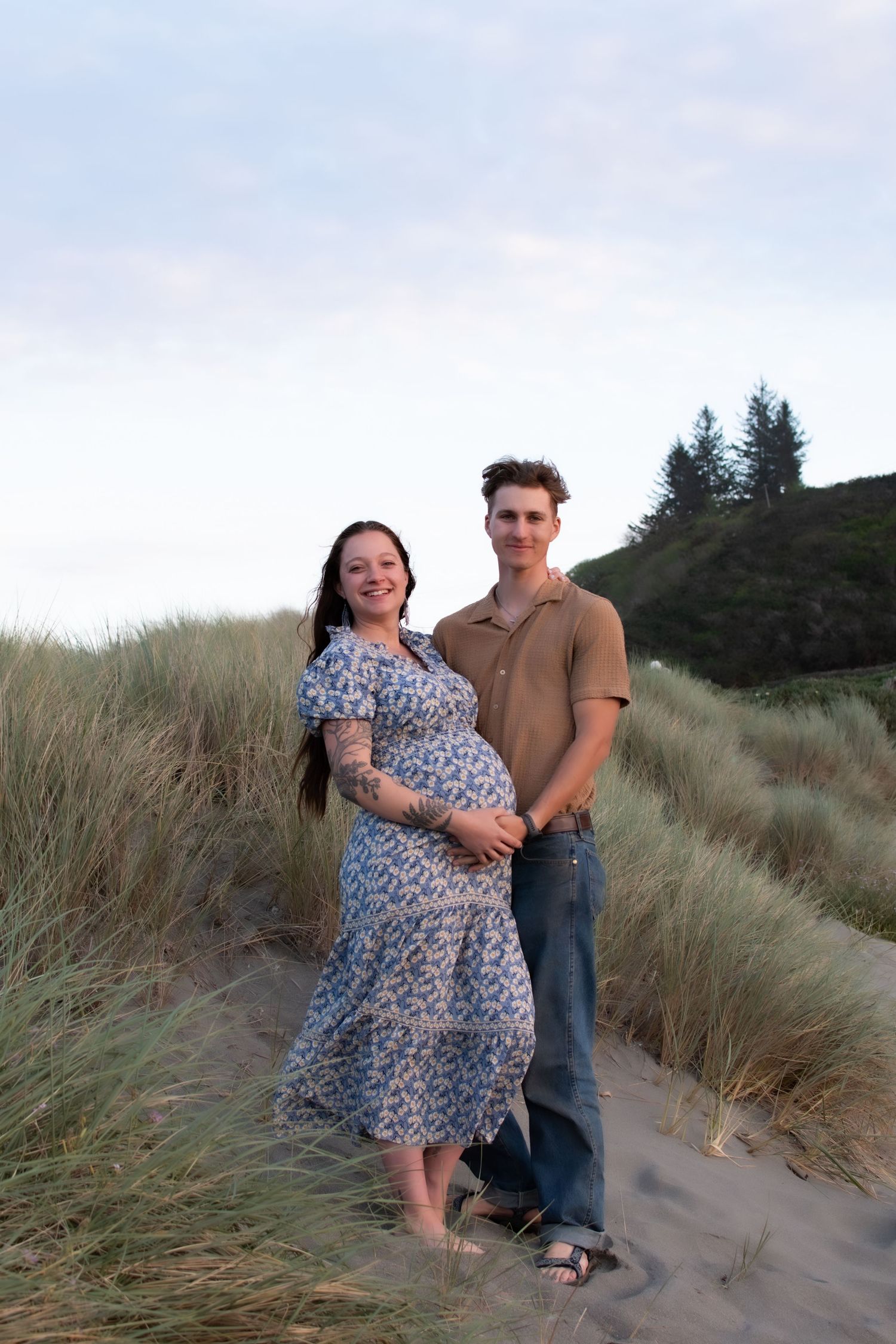 Pregnant woman in floral dress with partner on sandy beach dunes at dusk.