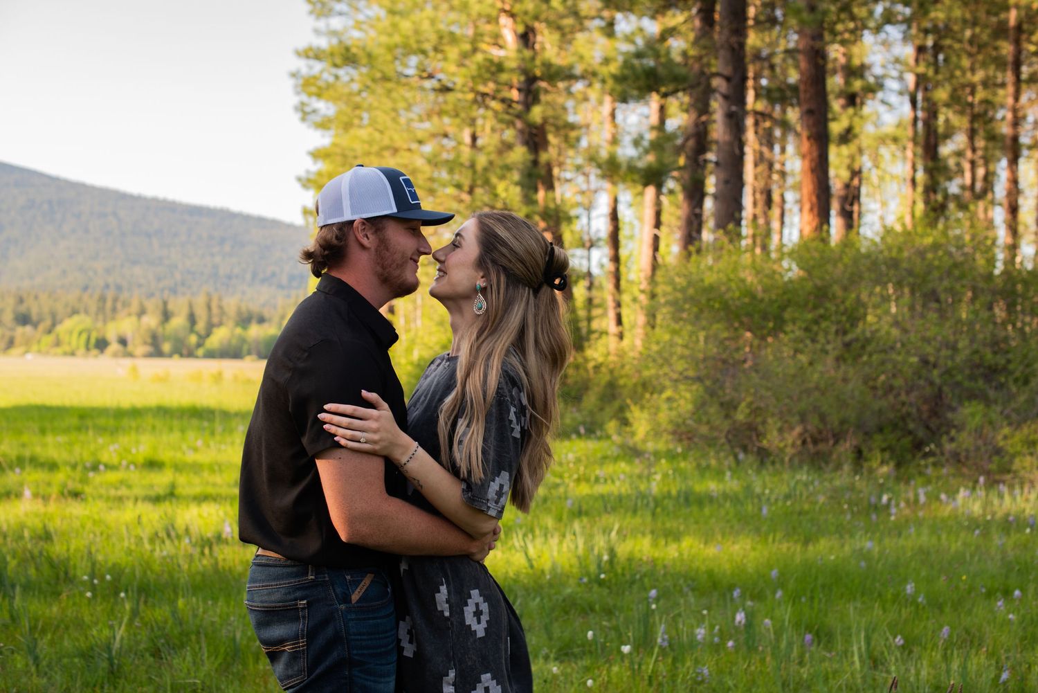 Loving couple embracing and about to kiss in a sunlit green meadow with pine trees and mountains.