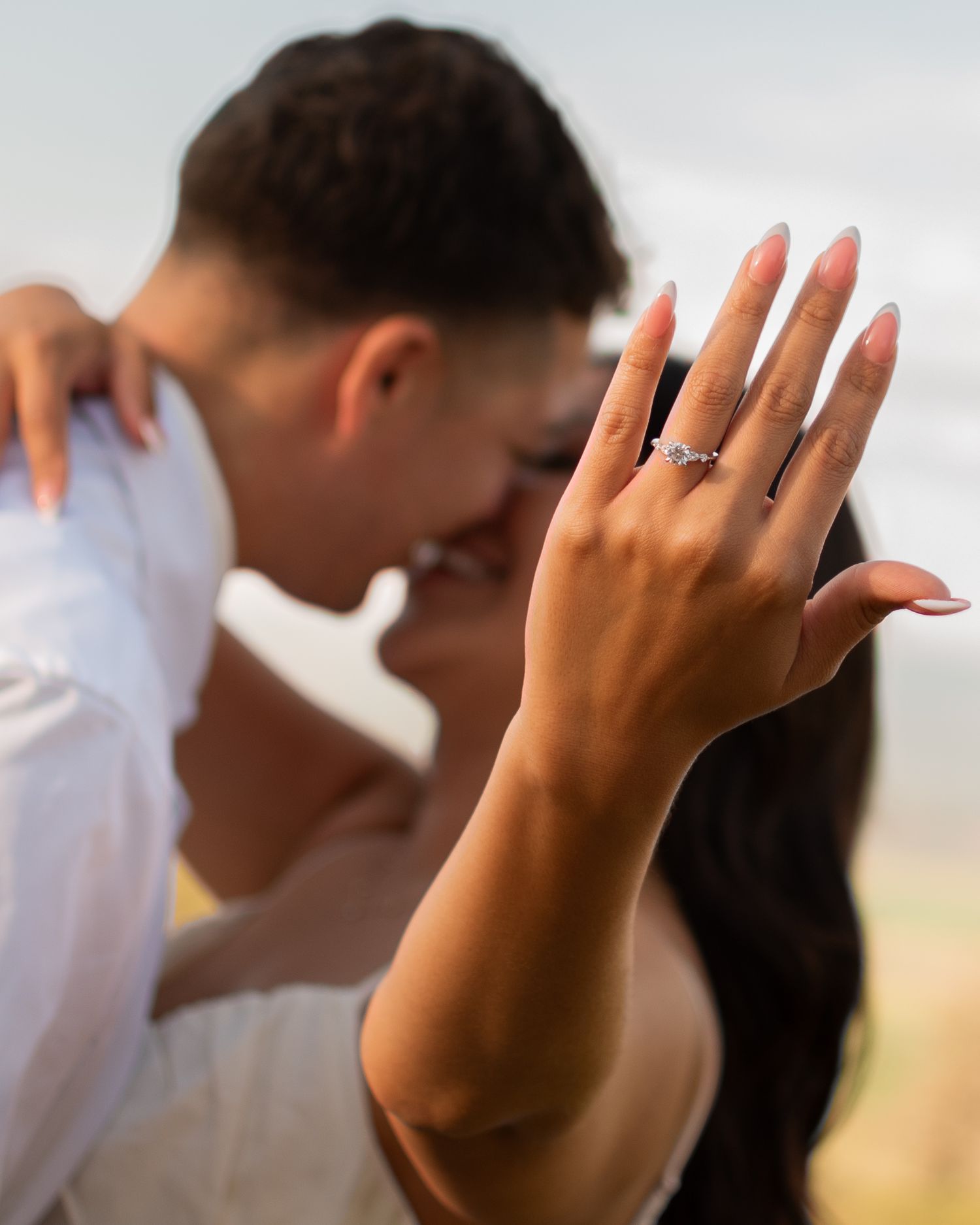 A couple shares a romantic kiss while the woman displays her diamond engagement ring in the foreground.