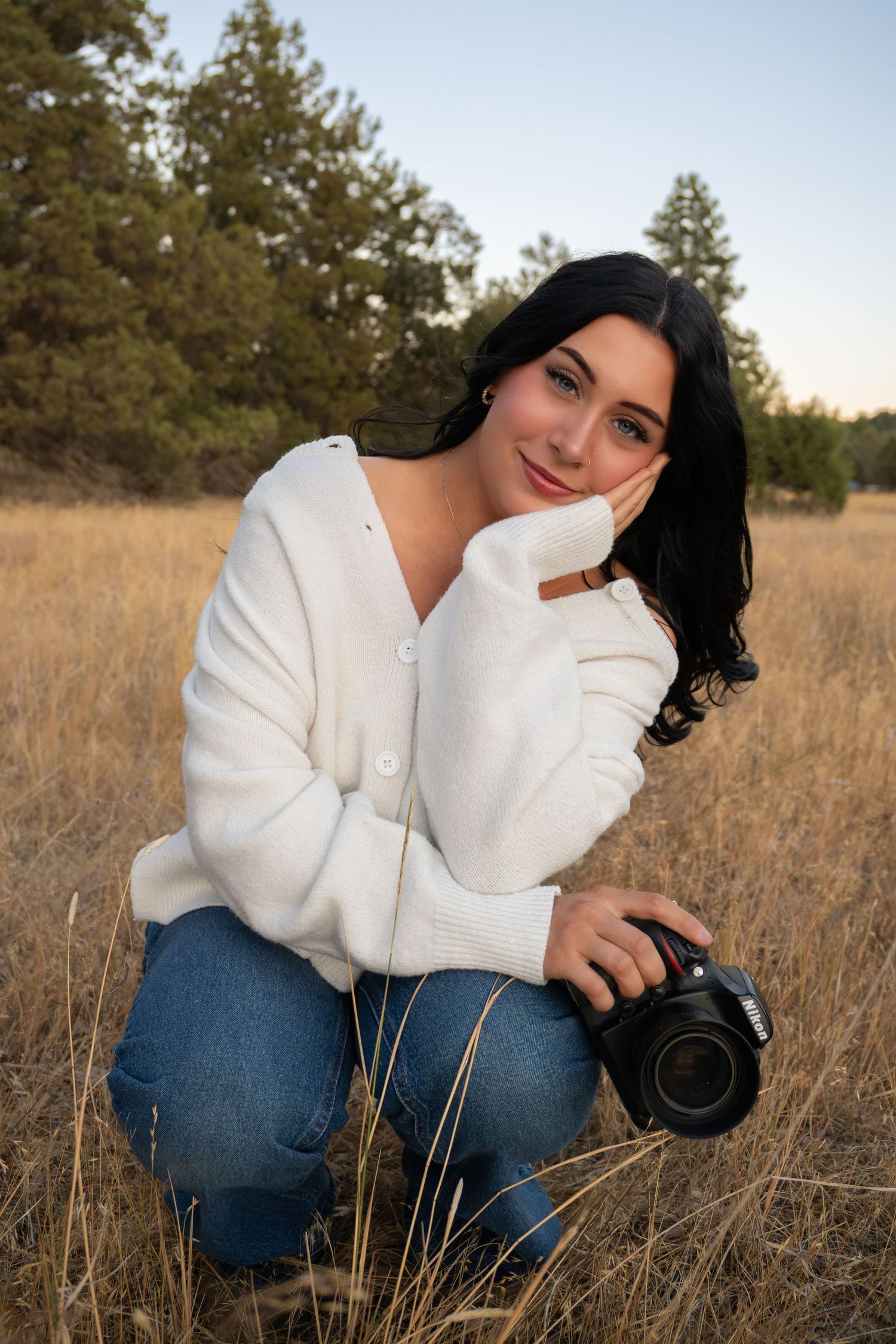 Young woman with dark hair holding a camera, crouching in a golden field wearing a white cardigan and jeans.