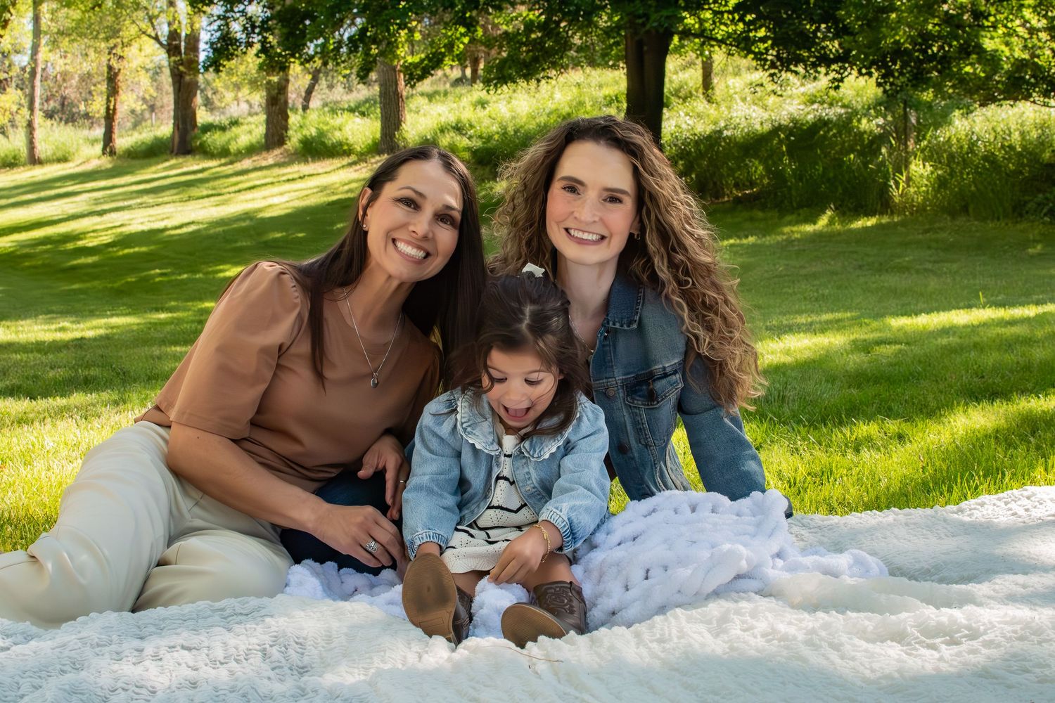 Two women and a young child smile together on a white blanket in a sunny green park setting.