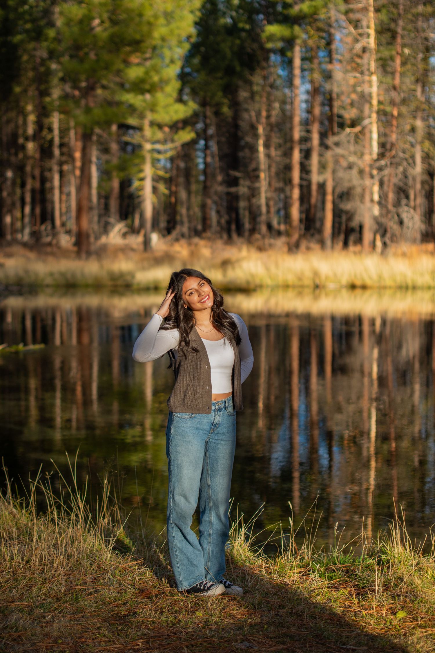 Young woman in jeans and brown jacket stands by a reflective forest pond in golden autumn light.