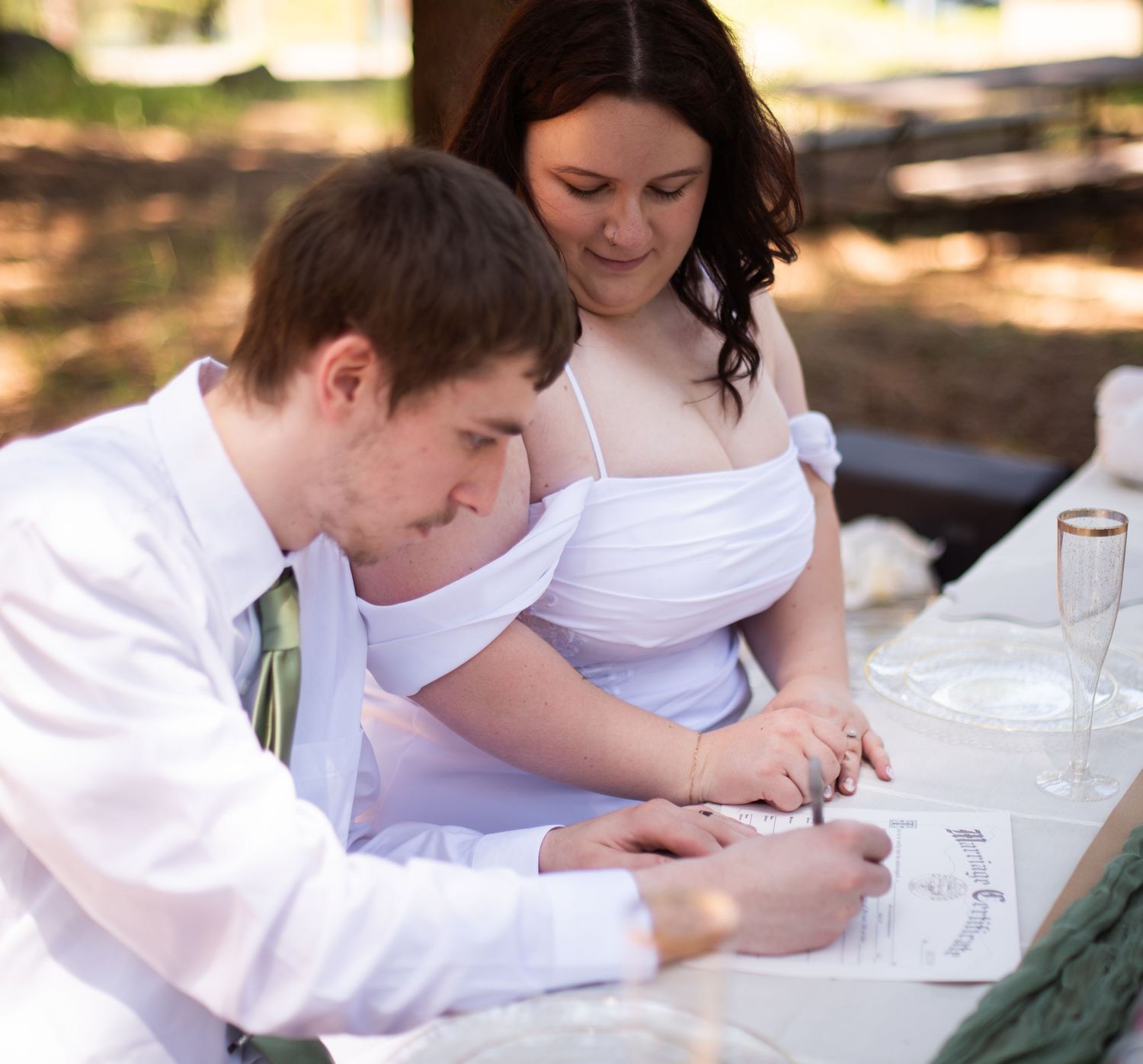 Newlyweds signing their marriage certificate together at an outdoor woodland wedding ceremony.