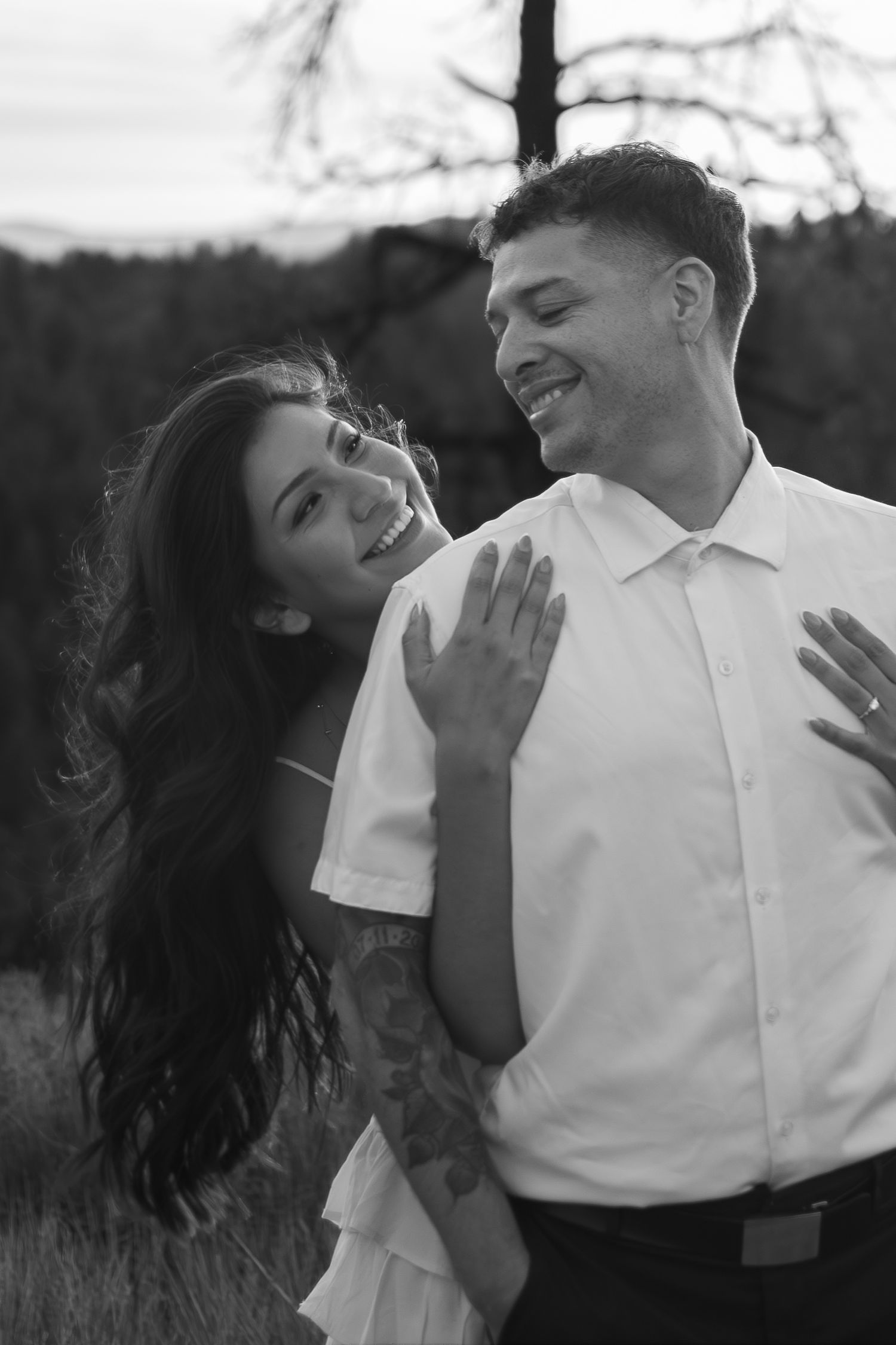 Black and white photo of a smiling couple embracing outdoors, man in white shirt with tattoos, woman in white dress.
