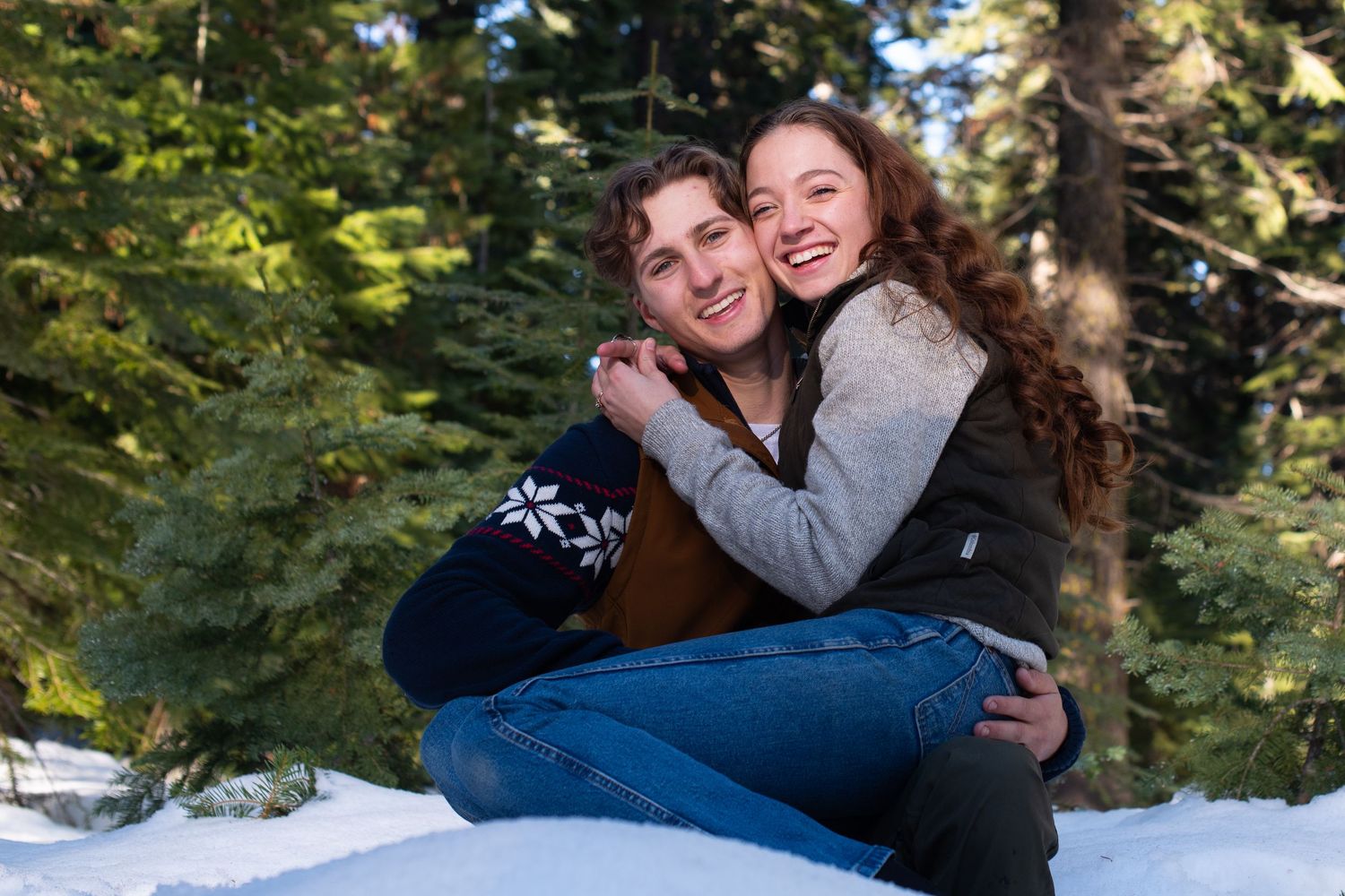 Two smiling women hugging while sitting in snow surrounded by pine trees.