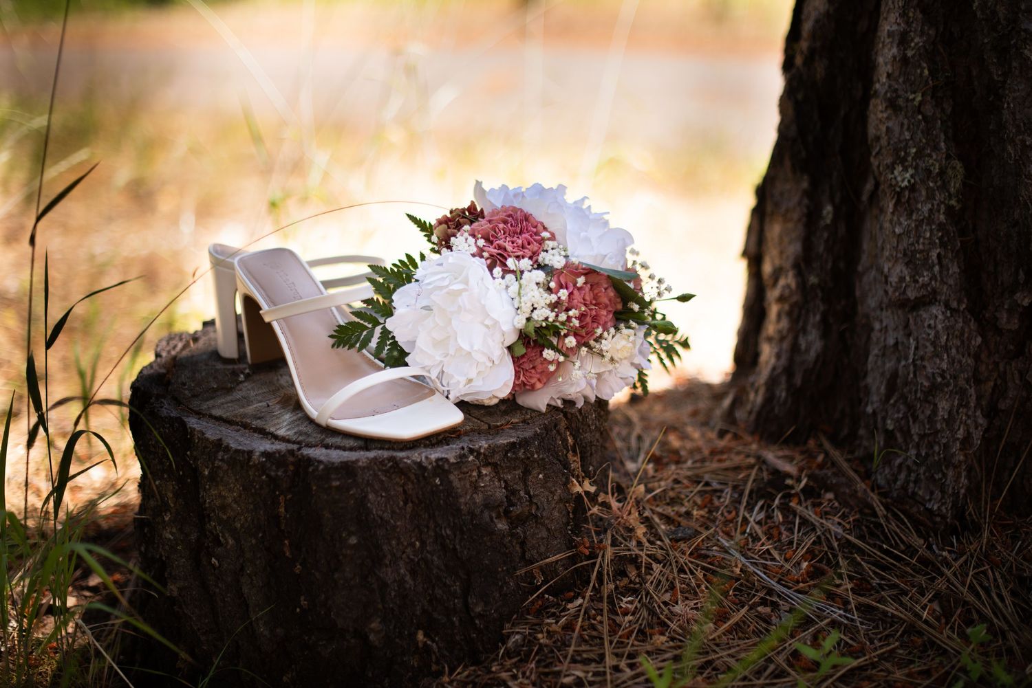 White bridal heels and a pink and white wedding bouquet displayed on a rustic tree stump outdoors.