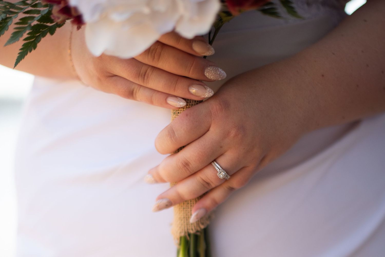 Close-up of bride's hands holding wedding bouquet with diamond engagement ring and manicured nails.