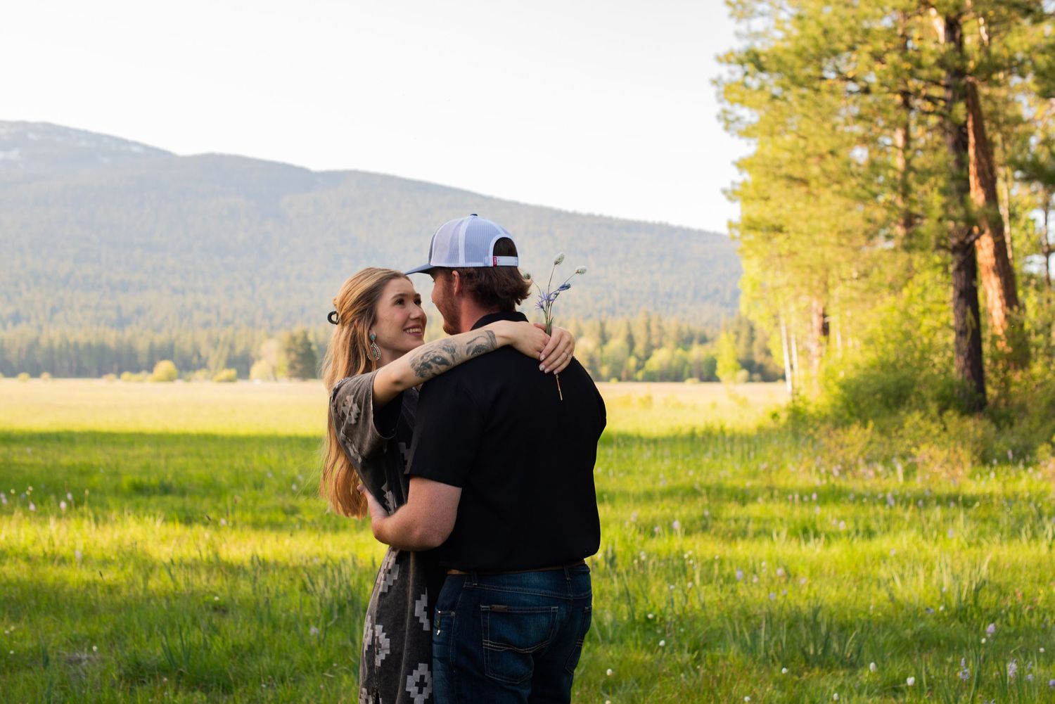 Romantic couple embracing in a green meadow with mountain views and pine trees at sunset.
