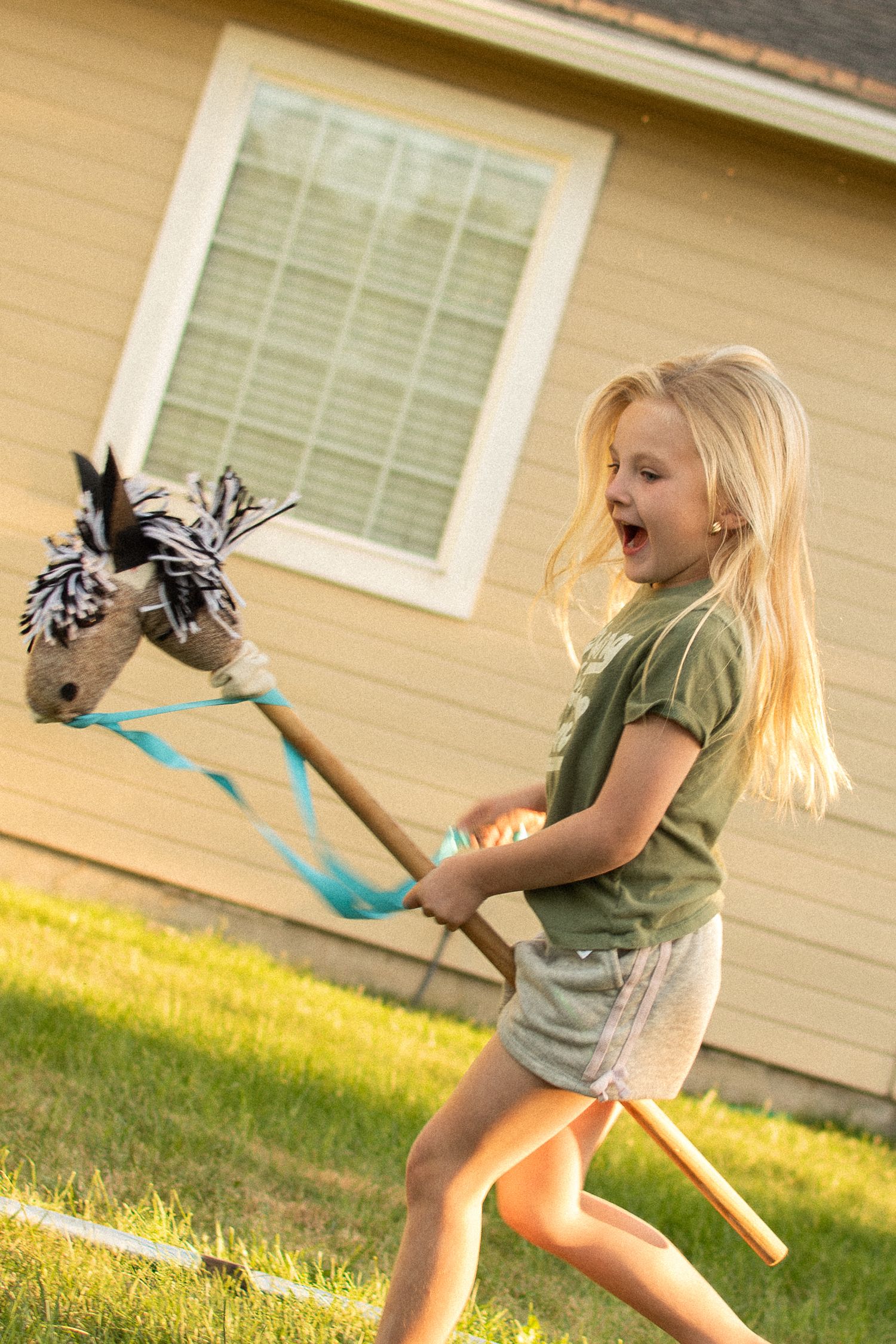 Joyful blonde girl running in backyard with hobby horse toy on sunny day.