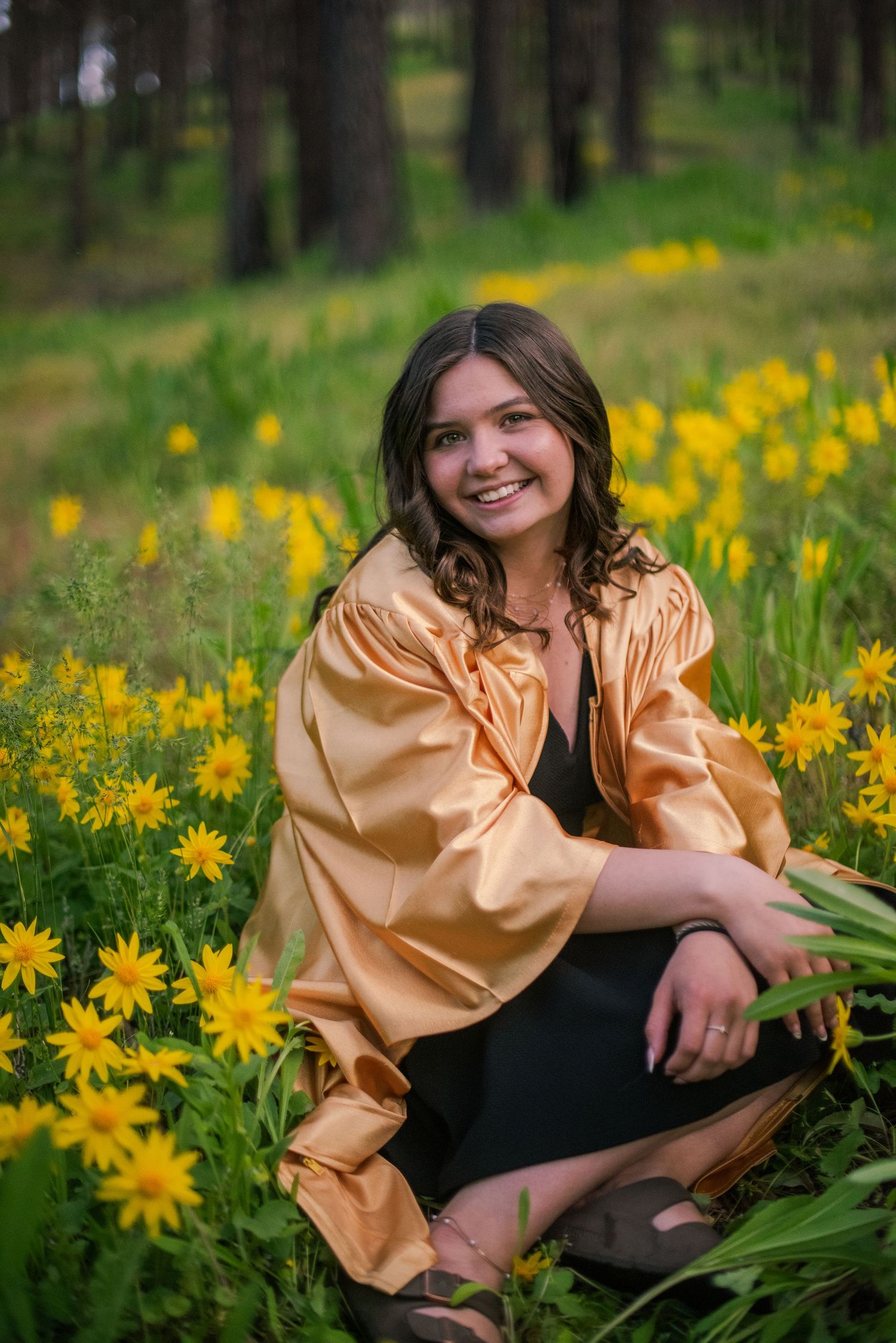 Smiling young woman in gold jacket sitting among bright yellow wildflowers in a pine forest.