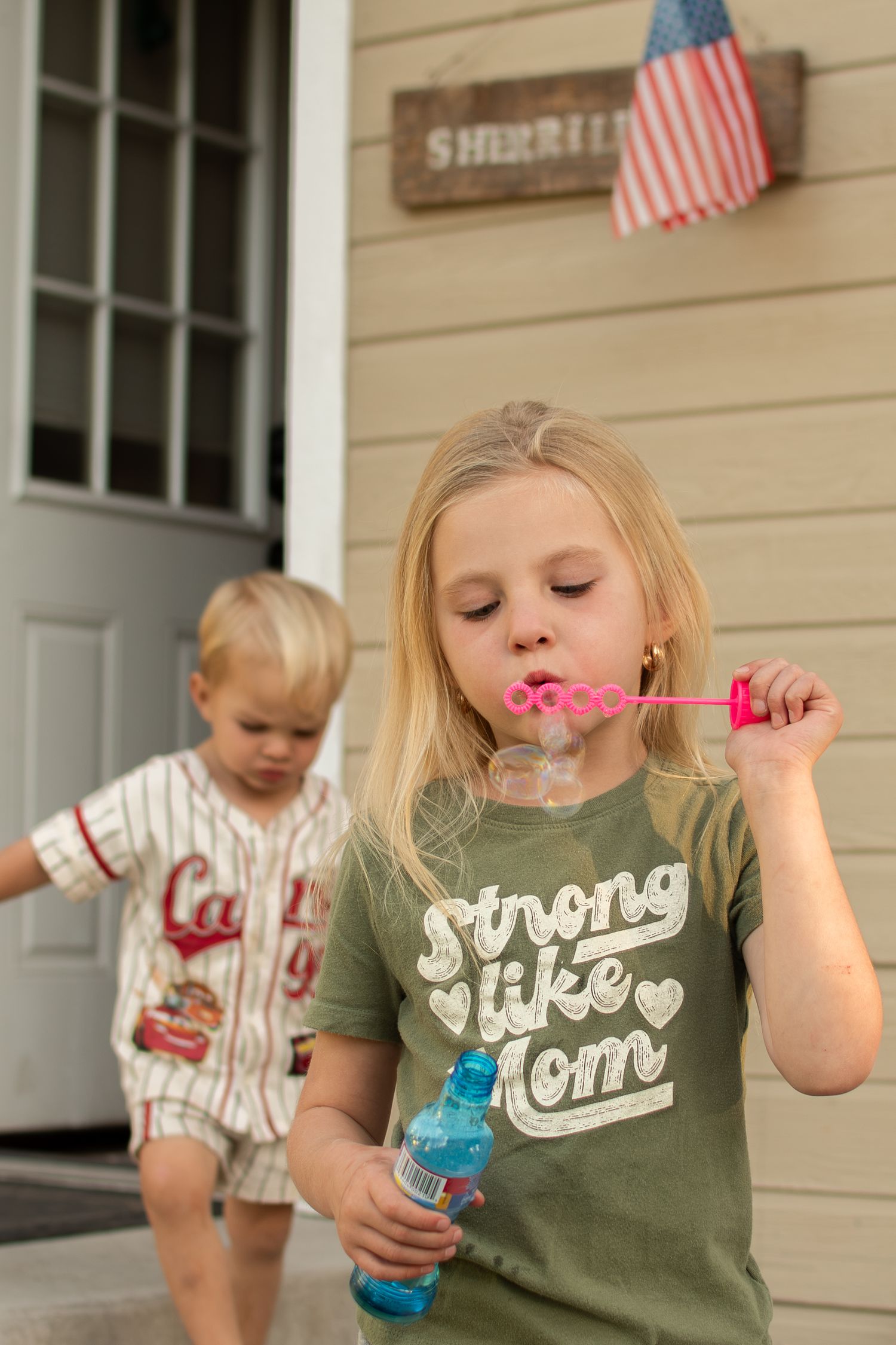 Girl in Strong Like Mom shirt blowing bubbles outside home with American flag, toddler boy in baseball uniform behind her.