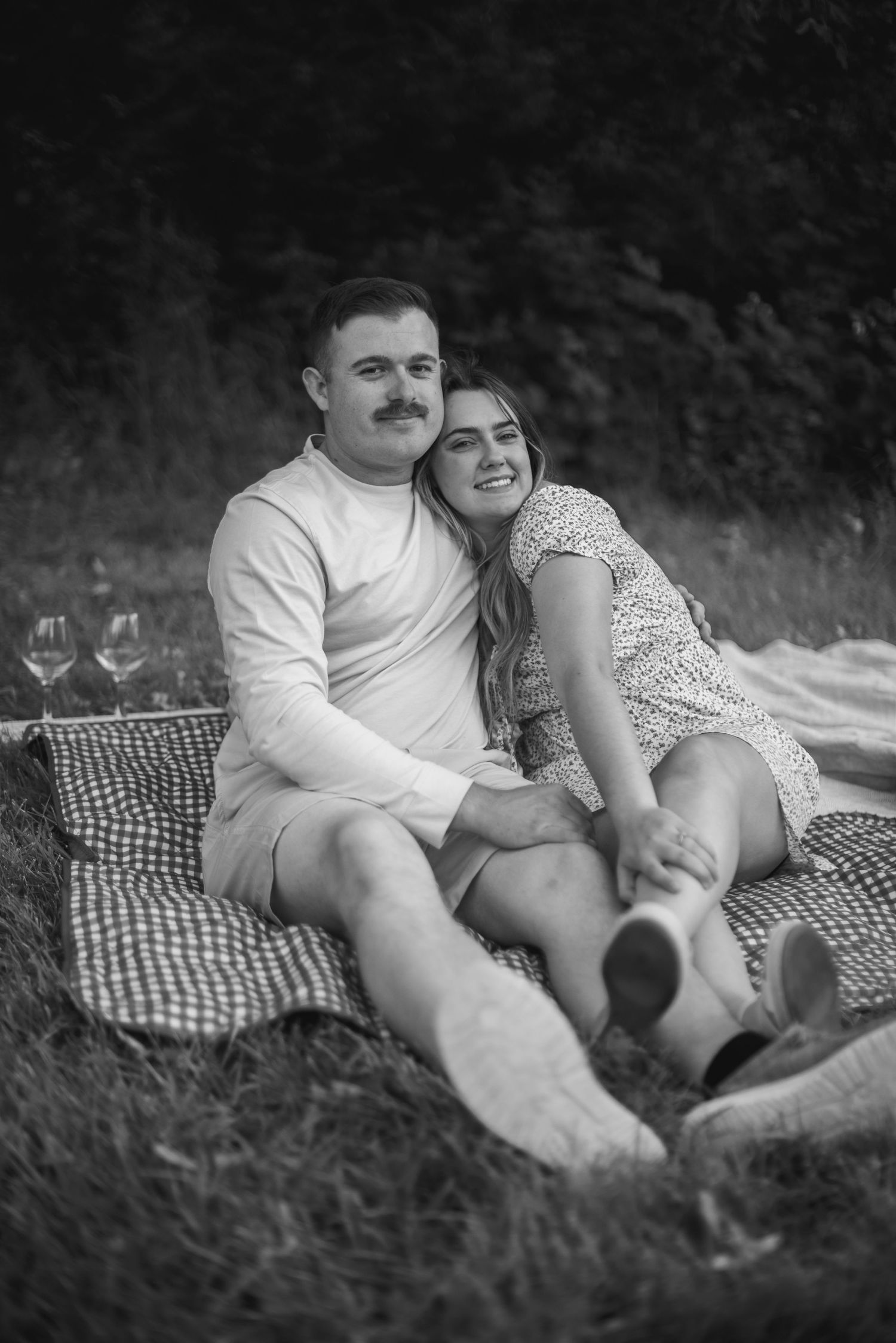 Black and white photo of a smiling couple sitting together on a picnic blanket outdoors.
