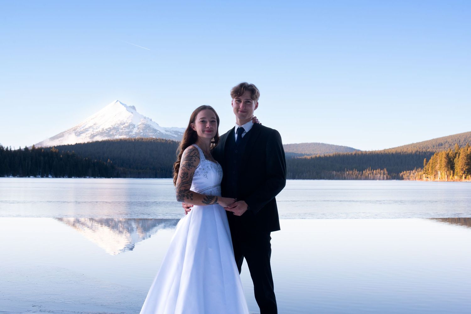 Bride and groom pose elegantly by a reflective mountain lake with a snow-capped peak backdrop.