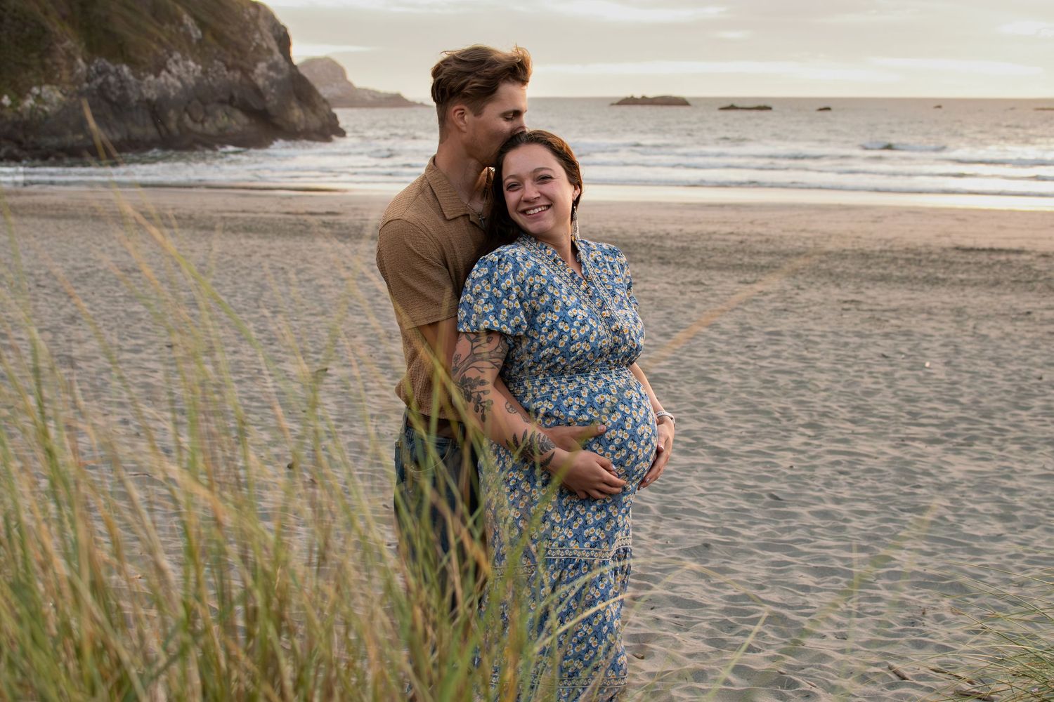 Couple embracing on a sandy beach at sunset, pregnant woman in floral dress smiling joyfully with partner behind her.
