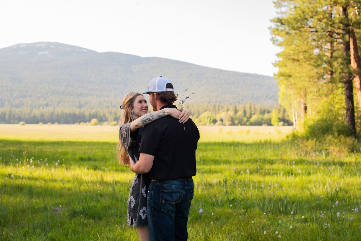 Couple embraces in lush green meadow with mountain backdrop.