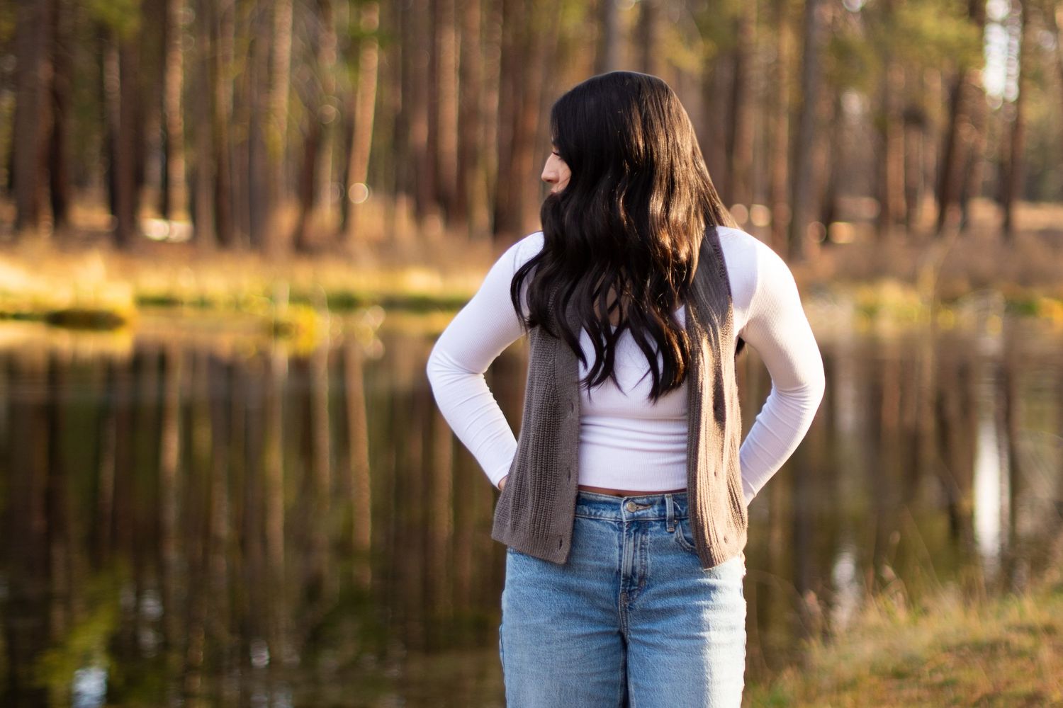Young woman with long dark hair in white crop top, vest, and jeans stands by a reflective forest lake.