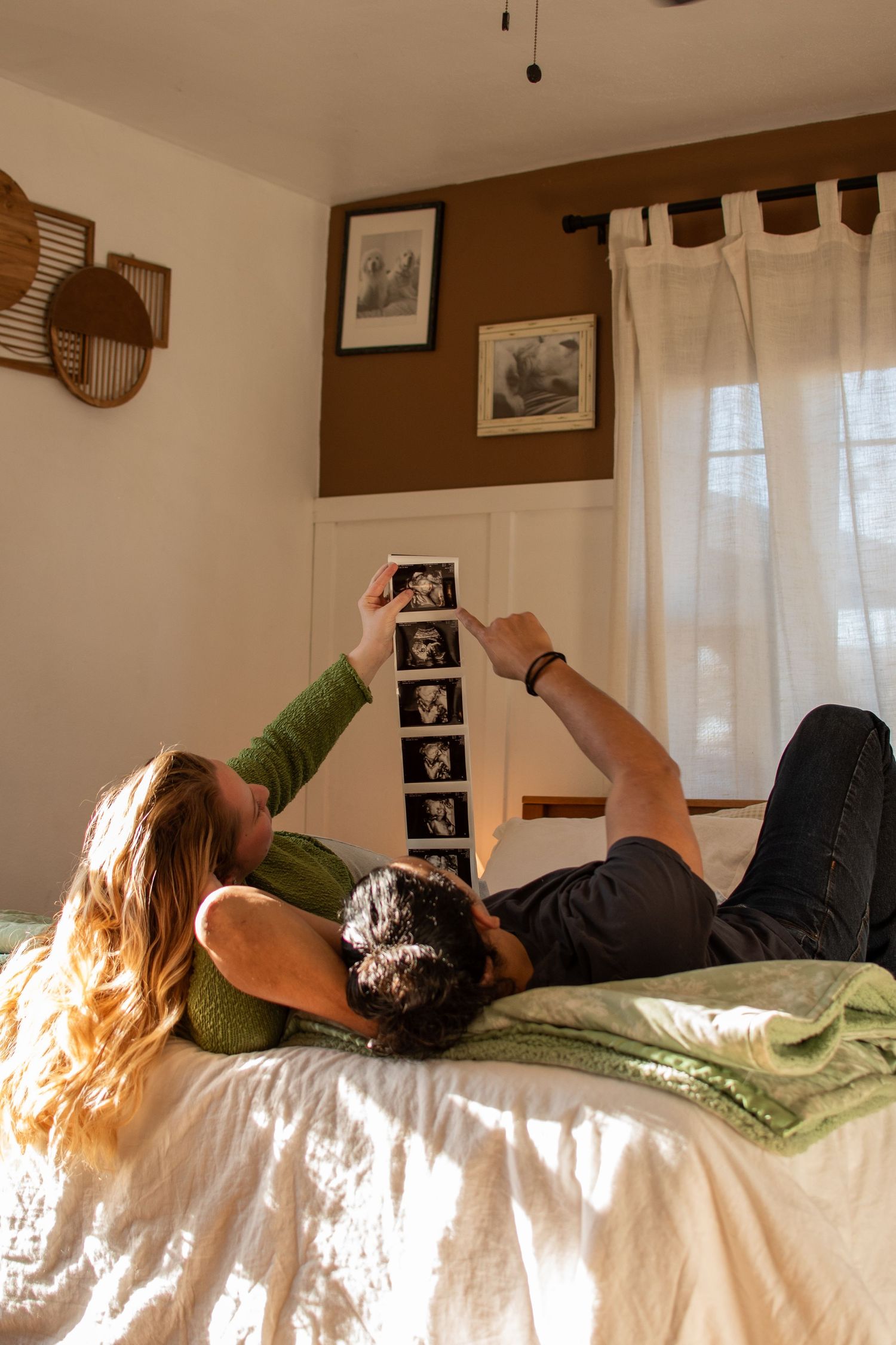 Couple lying on bed together holding up ultrasound photos in sunlit bedroom with brown walls and white curtains.