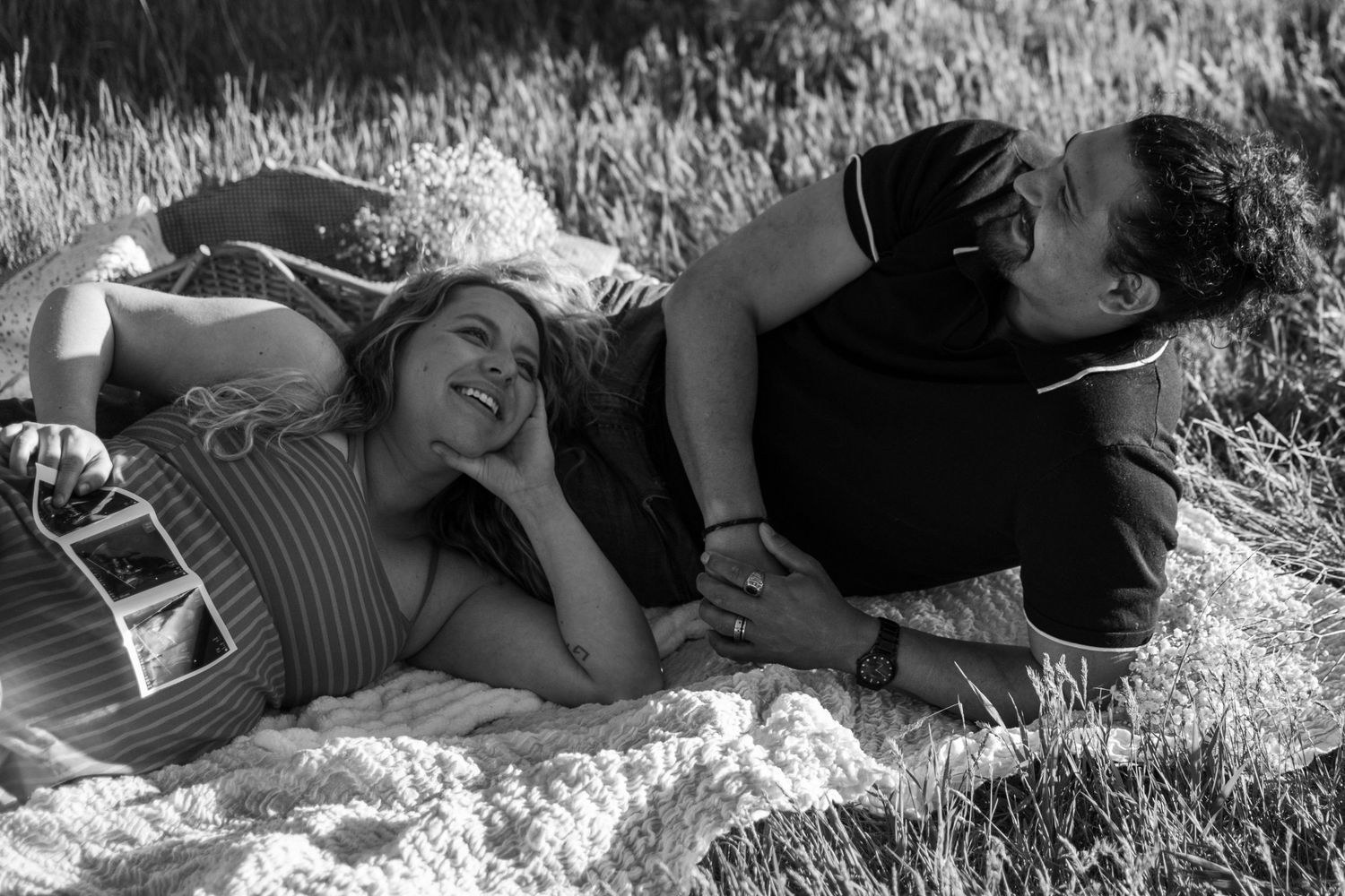 Black and white photo of a couple lying on a blanket in a field, smiling and gazing at each other.