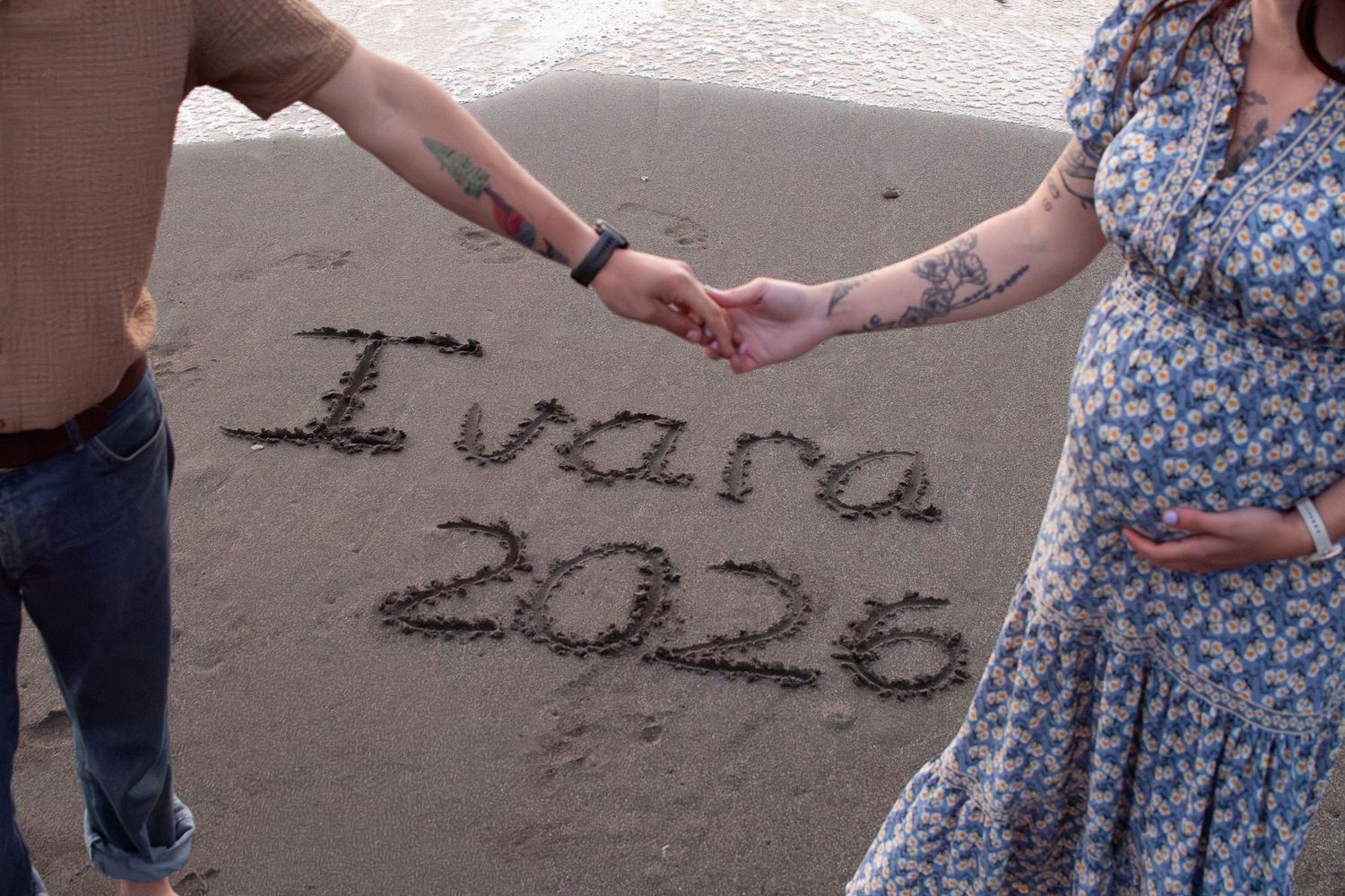 Couple holding hands over baby name Ivara 2026 written in sand on beach.