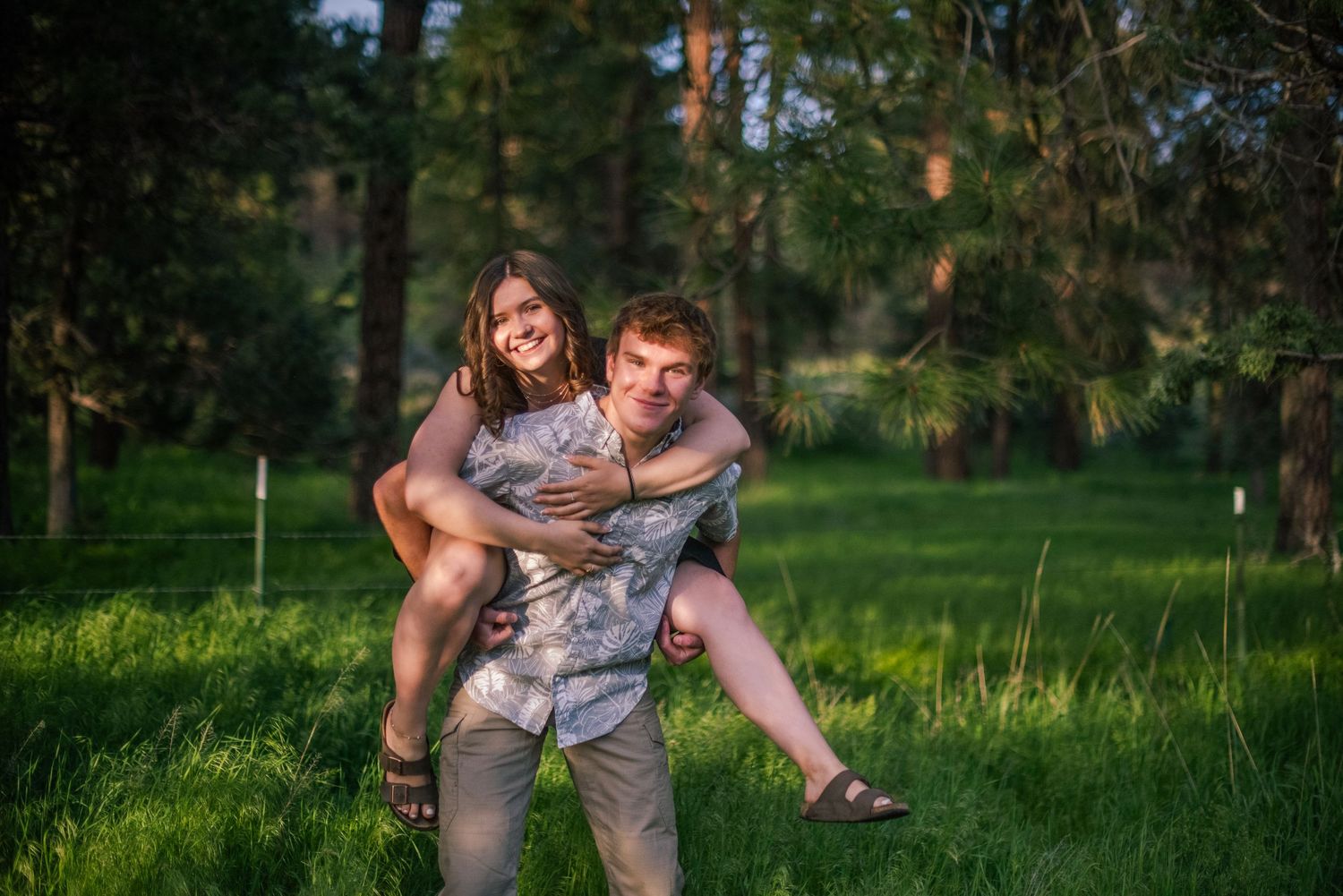 A young man gives a woman a piggyback ride while laughing in a sunny green forest clearing.